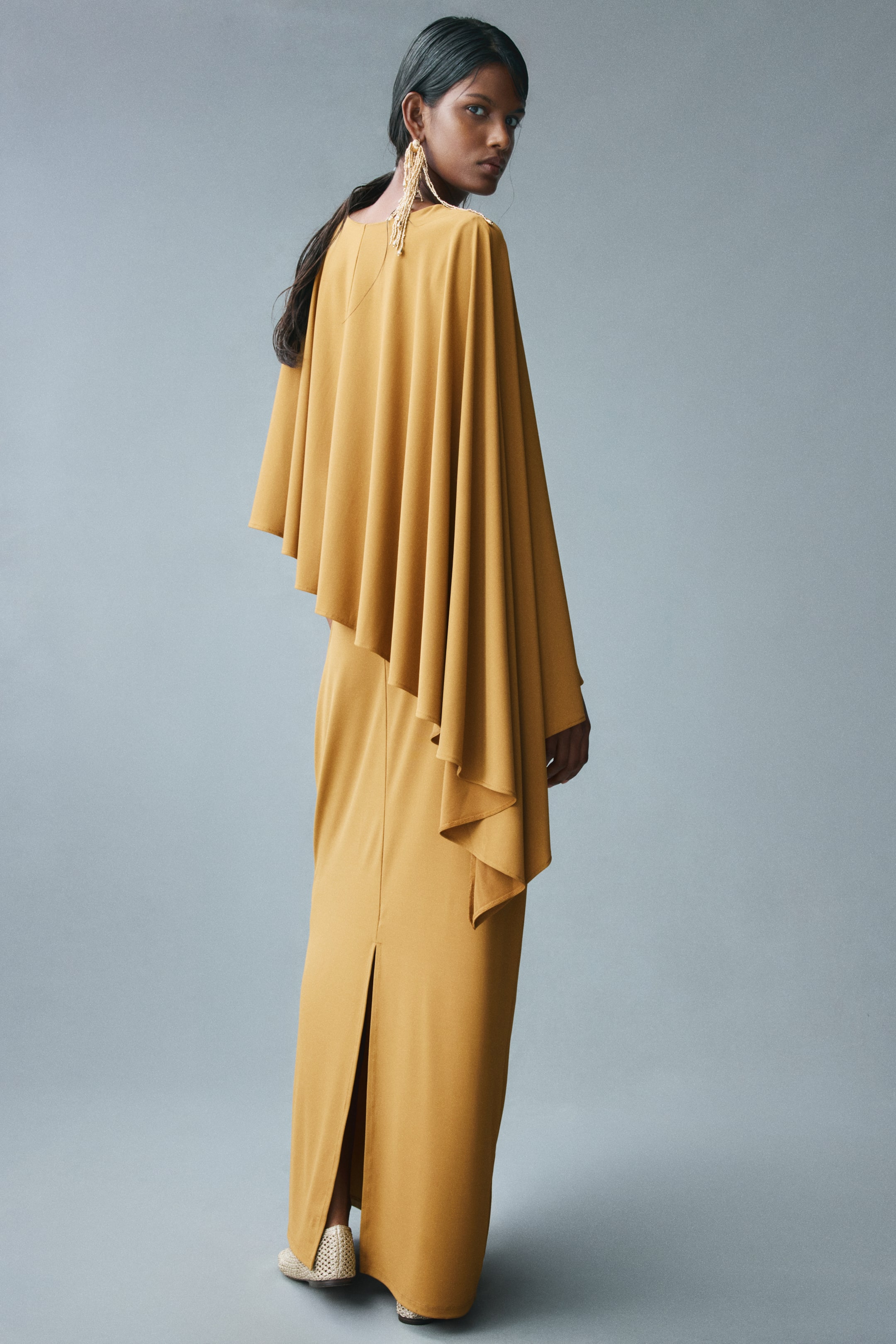 View larger image: A long mustard-yellow dress featuring a slim fit and a draped, asymmetrical cape-like layer is worn by the model. The dress has a straight-cut hem with a back slit, visible as the model stands and looks over their shoulder.