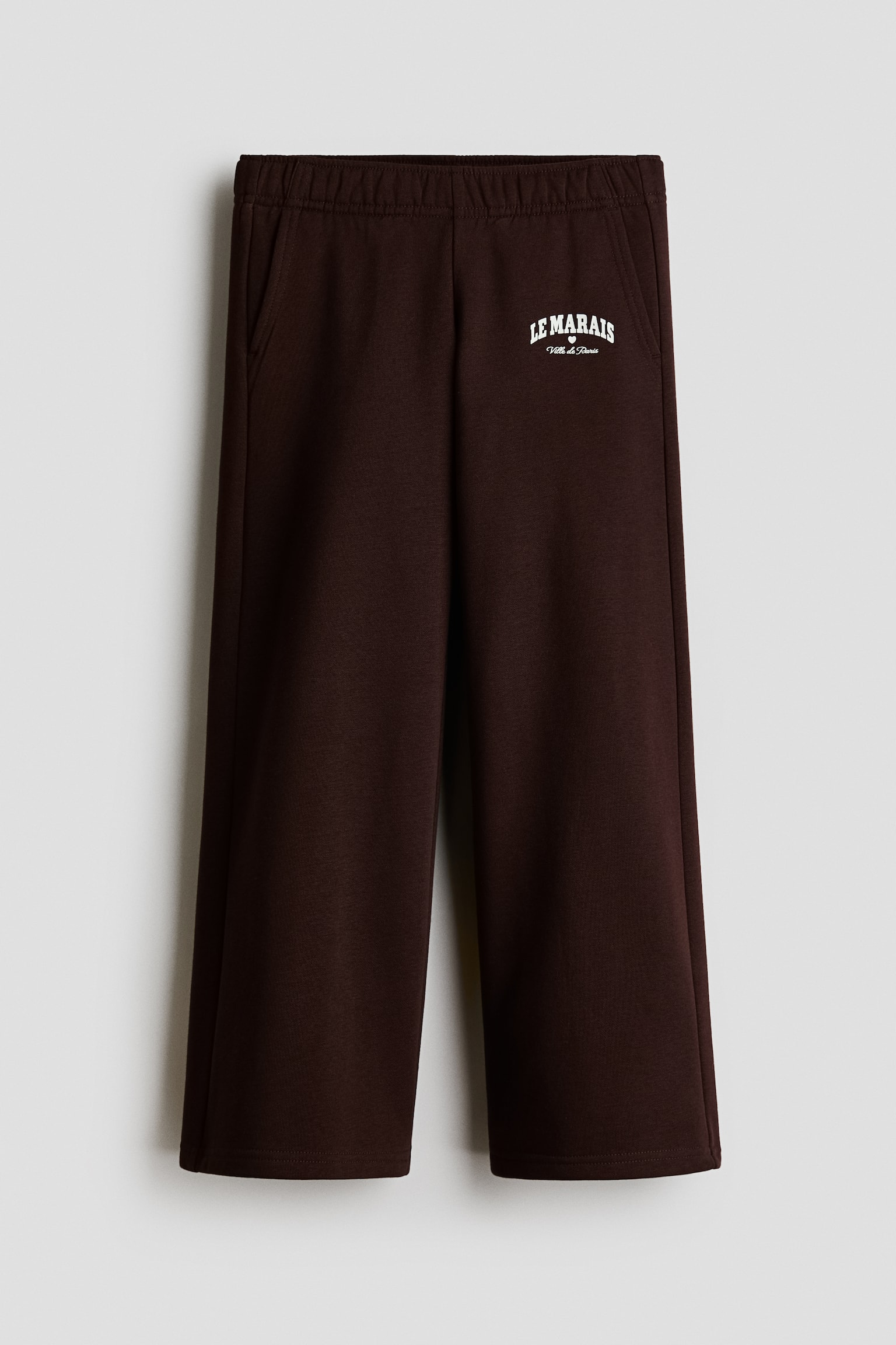 Donkerbruine joggingbroek van geborstelde fleecestof, van voren afgebeeld, met een elastische tailleband, schuine steekzakken en rechte pijpen. Op het bovenste deel van het linker bovenbeen staat een klein wit boogvormig grafisch detail met de tekst "LE MARAIS" boven "Ville de Paris", met in het midden een klein hartje.