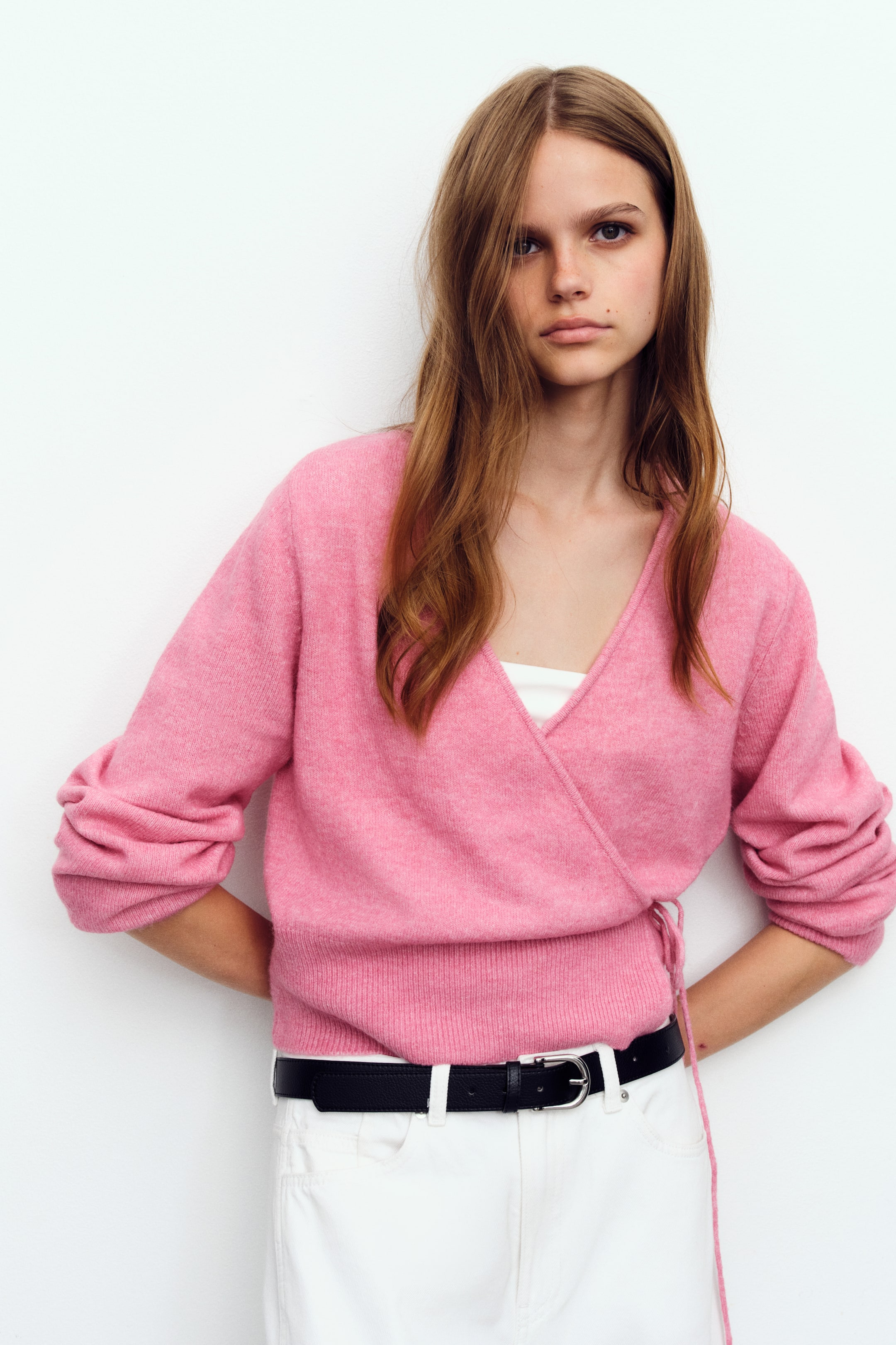 Προβολή μεγαλύτερης εικόνας: Ένα ροζ wrap cardigan από απαλό πλεκτό φοριέται από το μοντέλο, με λαιμόκοψη σε σχήμα V και μπροστινό μέρος wrap με λεπτά κορδόνια στερεωμένα στη μία πλευρά. Το cardigan φοριέται πάνω από λευκό τοπ και συνδυάζεται με λευκό παντελόνι και μαύρη ζώνη.