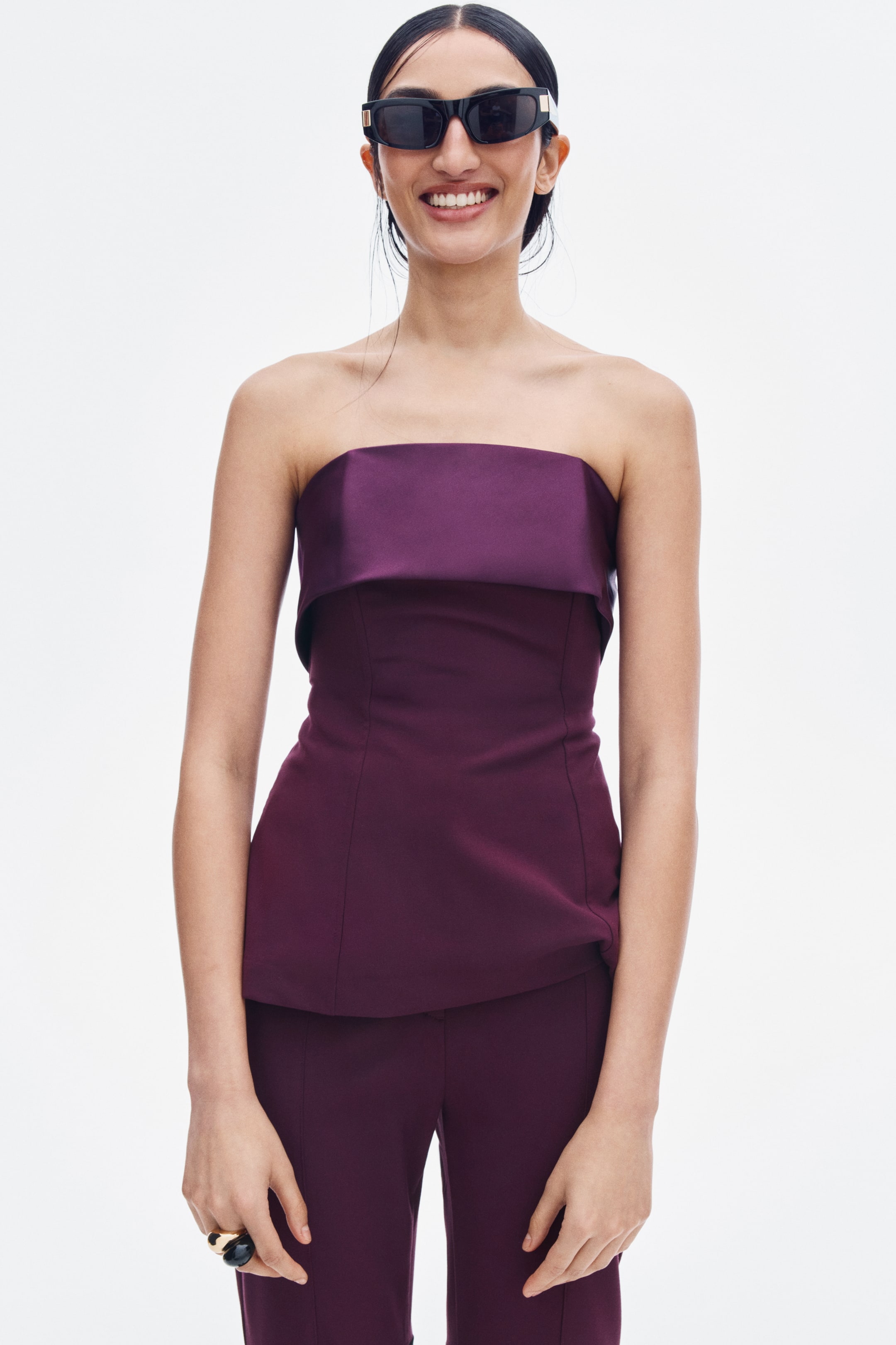 View larger image: A dark purple strapless top featuring a satin fold-over band at the bust and a structured, tailored bodice is being worn by the model. The top is styled with matching dark purple trousers.