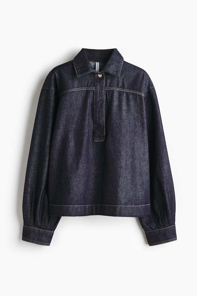 A dark wash denim shirt is shown FRONT, featuring a pointed collar and a partial button placket with a single gold-toned button. Contrasting tan topstitching is detailing the seams, and the long sleeves are gathered above the buttoned cuffs. - 1
