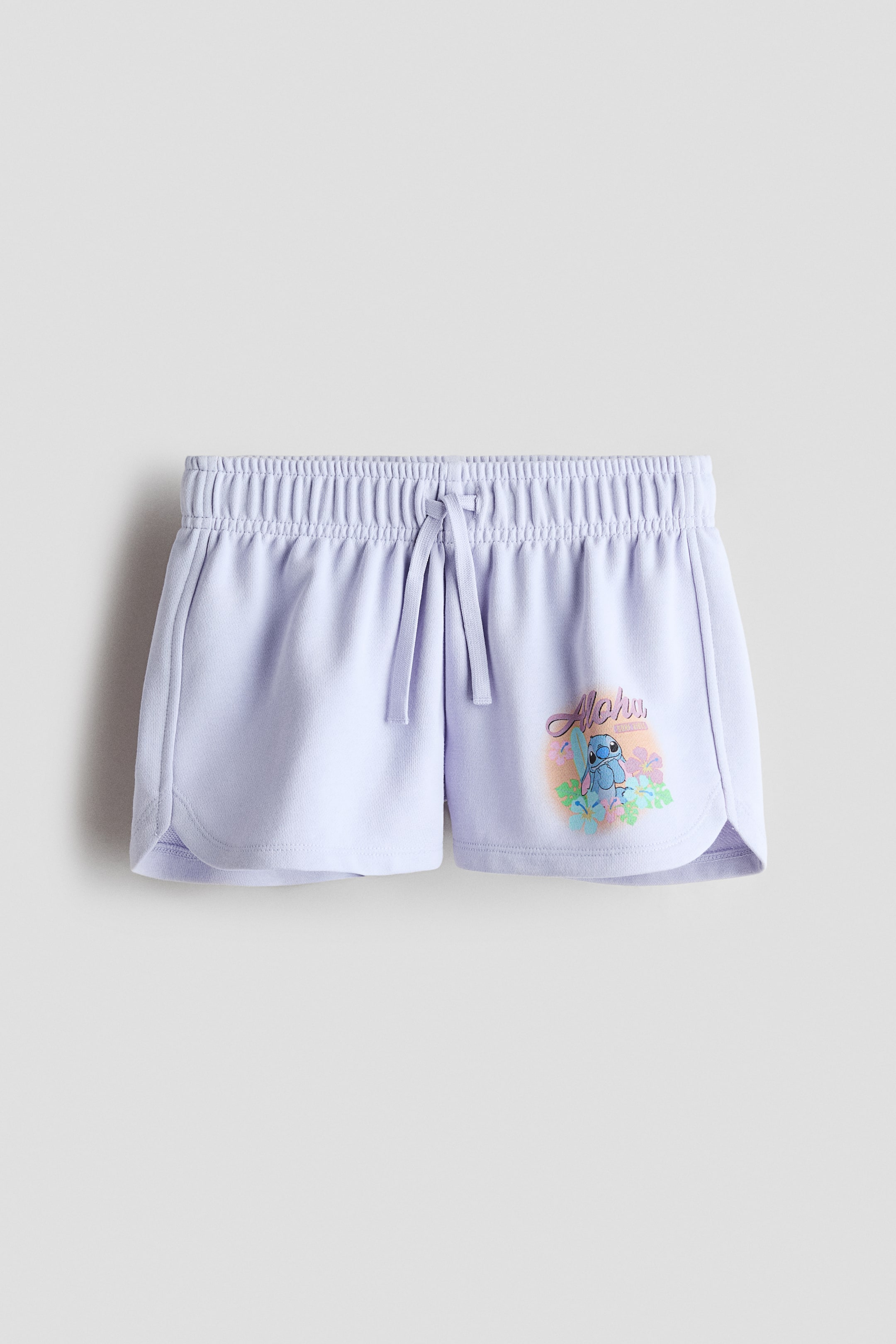 Grösseres Bild ansehen: Hellviolette Shorts zeigen eine Vorderansicht und verfügen über einen elastischen, gerafften Bund mit gebundenem Kordelzug. Weiße Paspeln verzieren die Seitennähte und die abgerundeten Säume, während ein farbenfrohes "Aloha"-Grafikdesign und die Figur Stitch zwischen tropischen Blumen das rechte Bein schmücken.