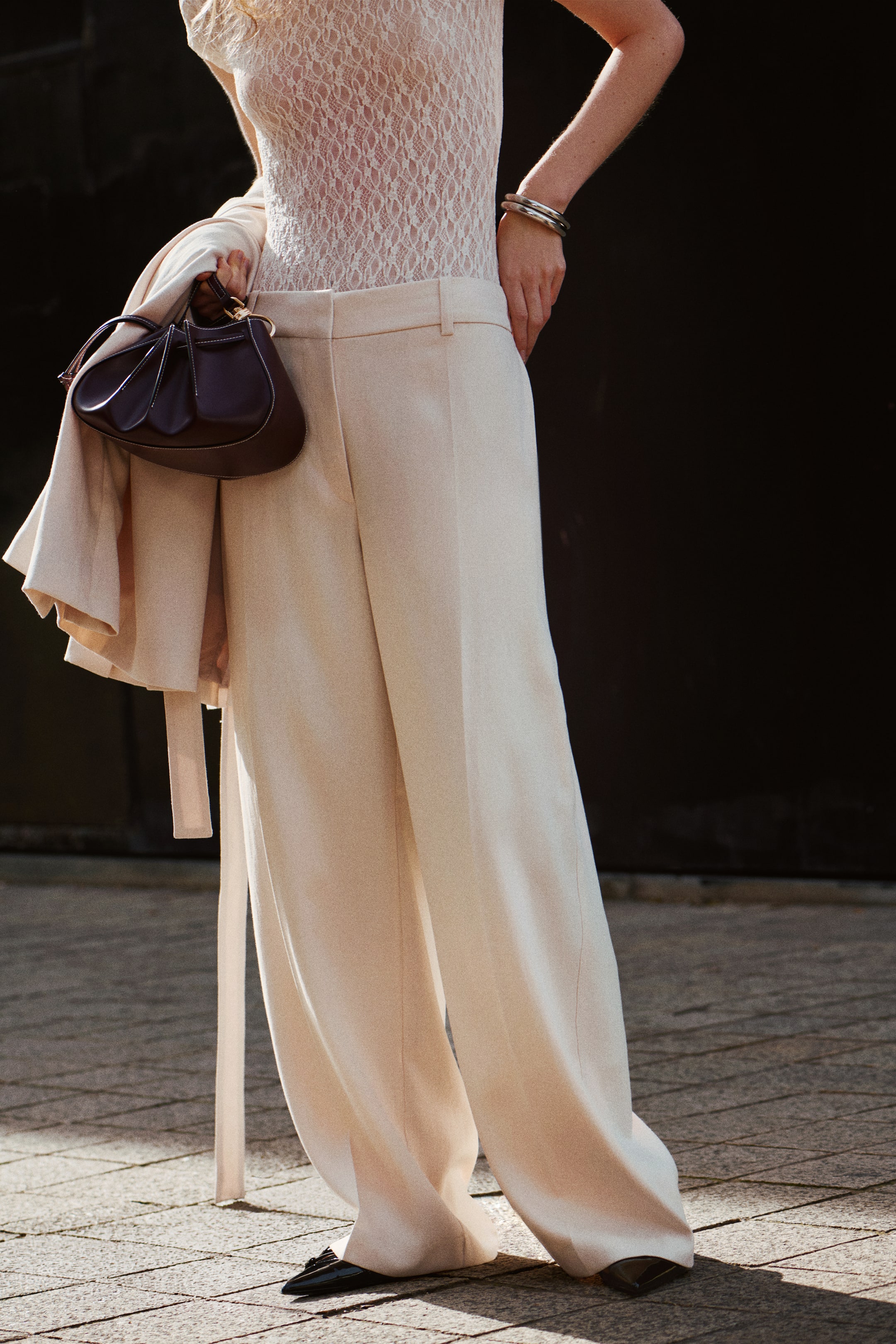 View larger image: Cream wide-leg trousers are displayed from a slight angle, showcasing a high-waisted, tailored fit. The trousers drape smoothly, with a wide silhouette that reaches the ground and partially conceals black flat shoes.