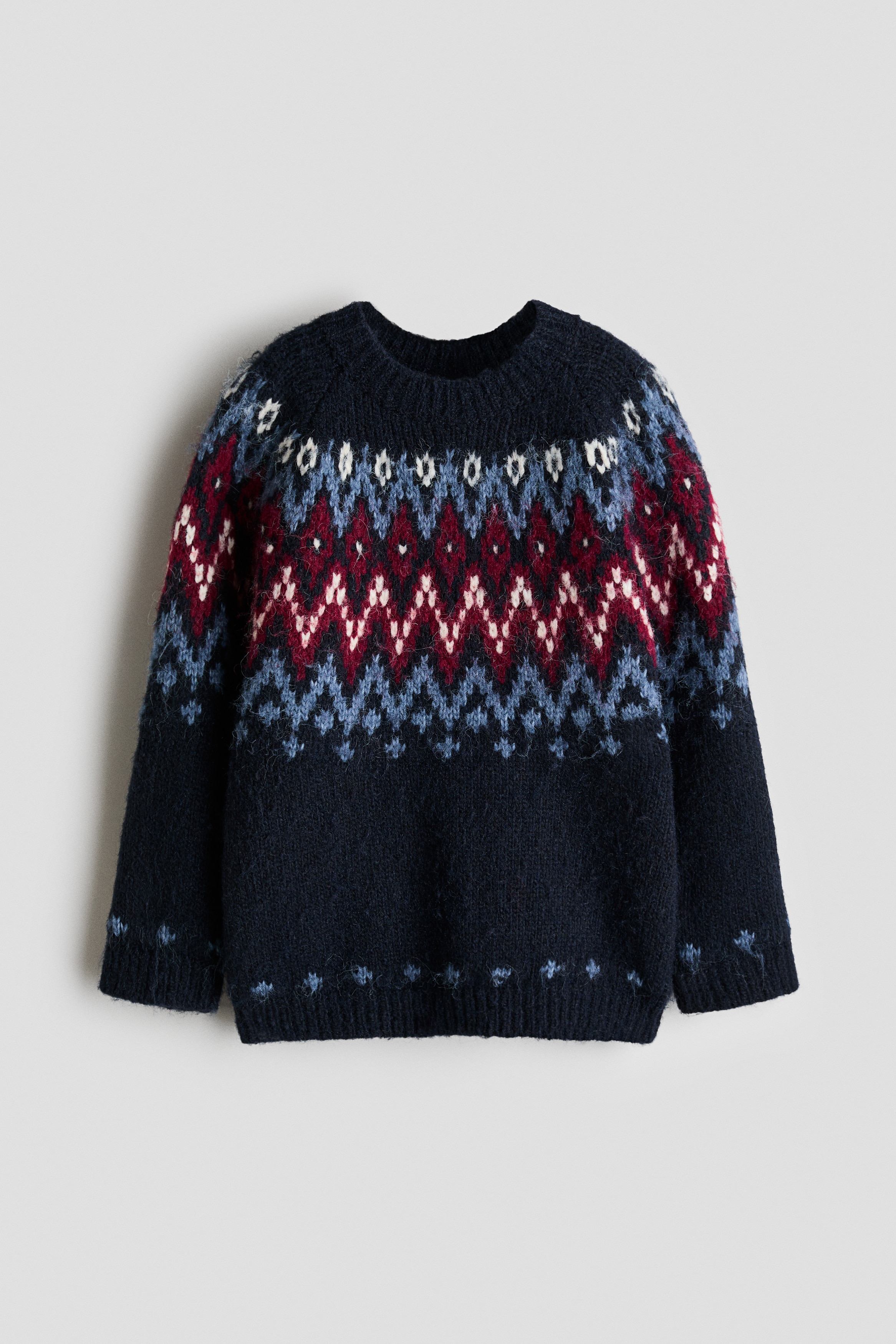A loose-fit sweater in a soft, navy blue jacquard knit is shown from the front, featuring a round, rib-trimmed neckline and long raglan sleeves. The sweater showcases a geometric knit pattern in shades of blue, burgundy, and white across the upper chest and shoulders, and is finished with ribbed cuffs and a ribbed hem.