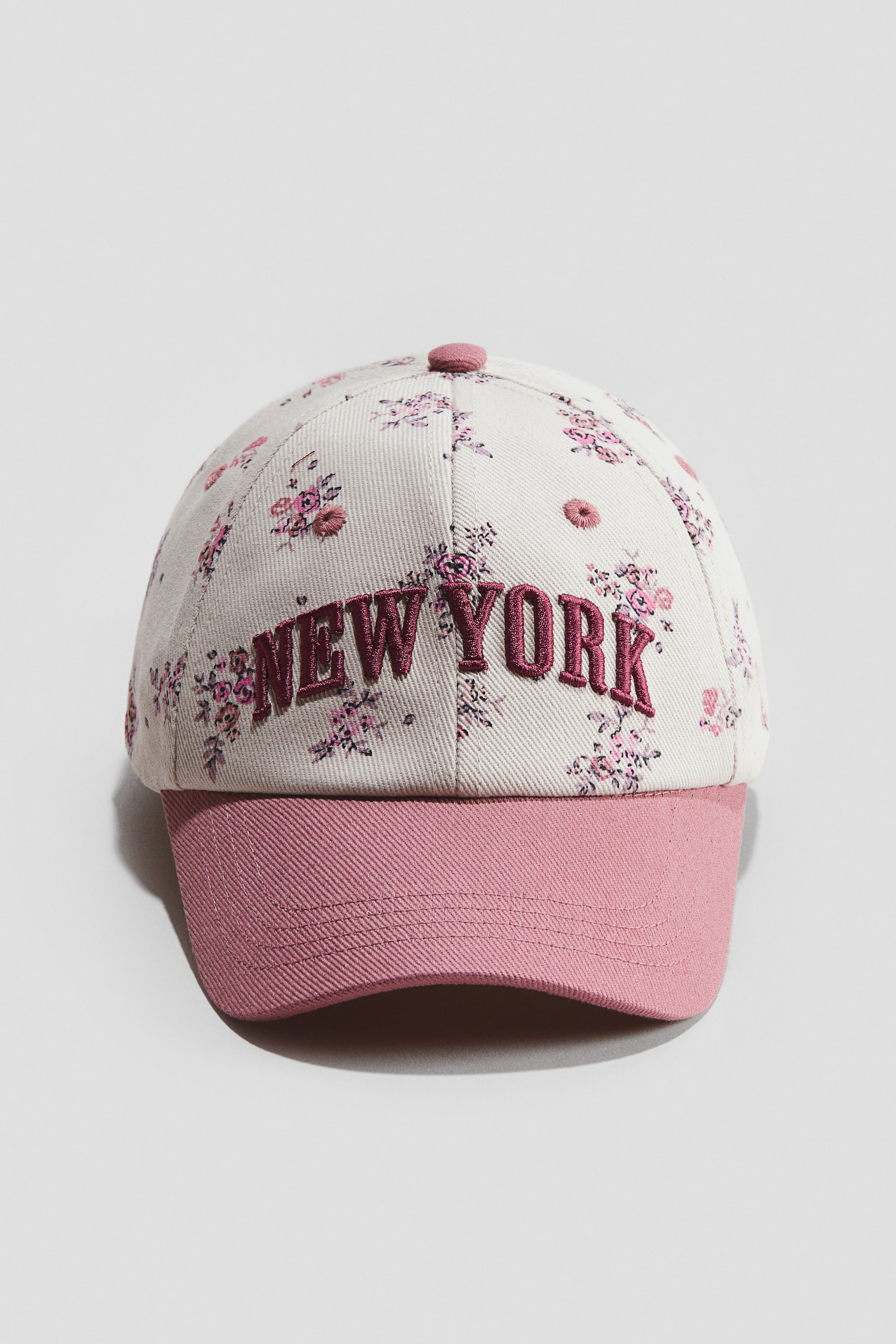 Voir une image plus grande: Une casquette de baseball présente une couronne blanc cassé ornée de motifs floraux roses et violets, avec une visière rose unie. Le devant de la couronne affiche "NEW YORK" brodé en lettres roses foncées en relief.