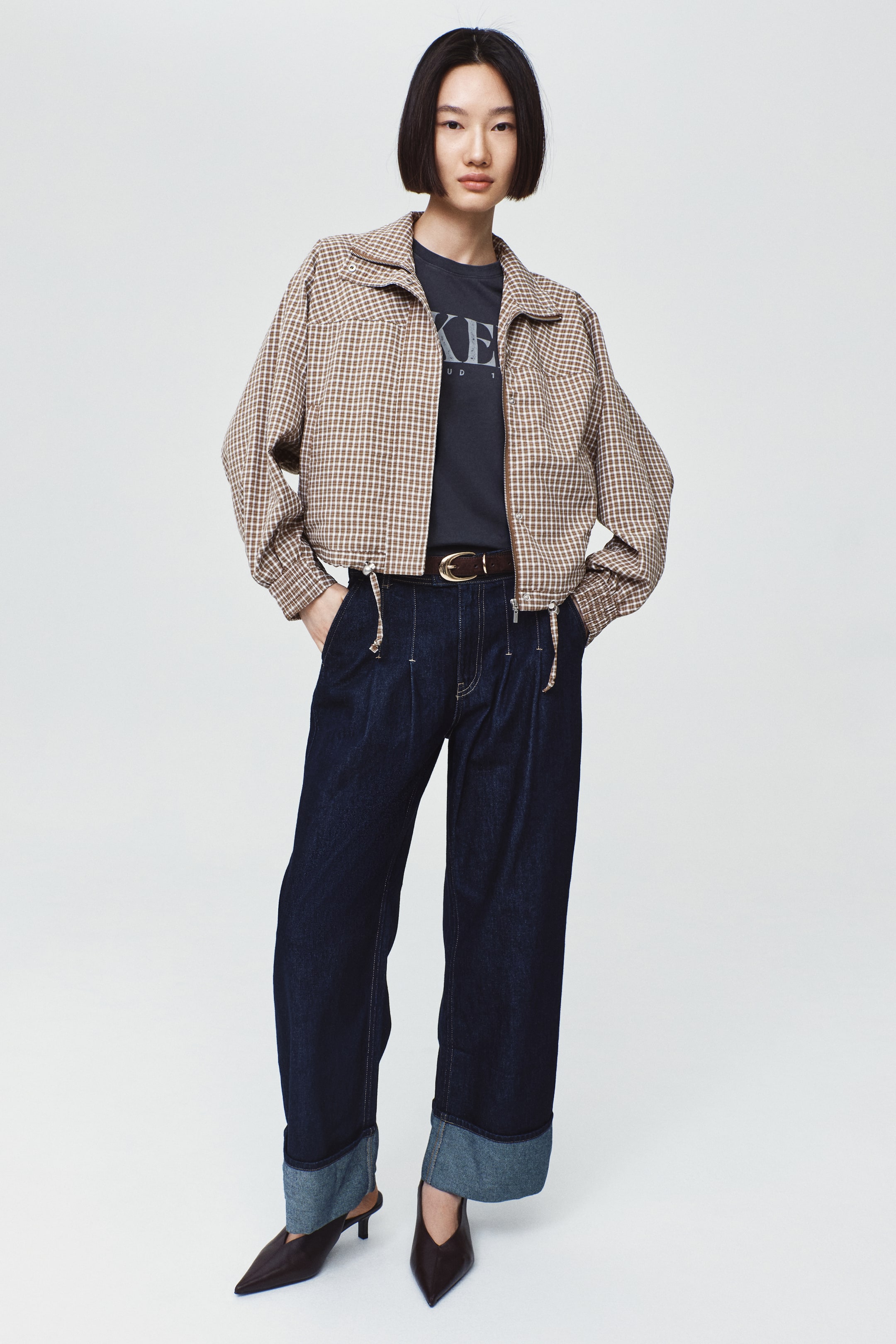 View larger image: A brown-and-white checked jacket with a textured weave is worn open by the model, featuring a stand collar, long dolman sleeves, and a drawstring hem with visible cord stoppers. The jacket is layered over a dark grey graphic T-shirt and paired with dark, wide-leg denim jeans.