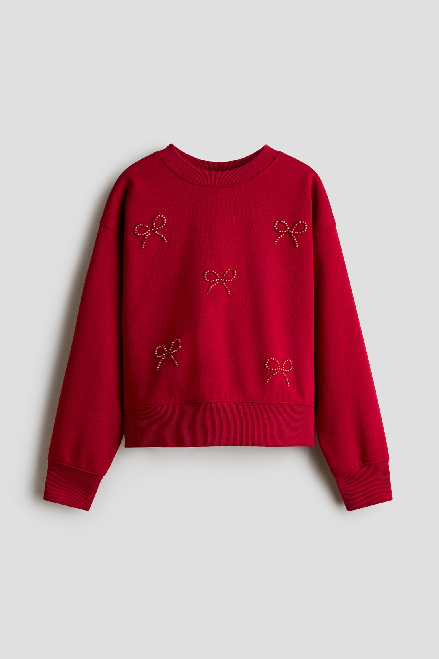 Un chandail Ã  coupe ample rouge est prÃ©sentÃ© de face, mettant en valeur un col rond bordÃ© de cÃ´tes, des Ã©paules tombantes, ainsi que des poignets et un ourlet cÃ´telÃ©s. Cinq petits motifs de nÅuds en perles rouges scintillantes ornent le devant du vÃªtement.