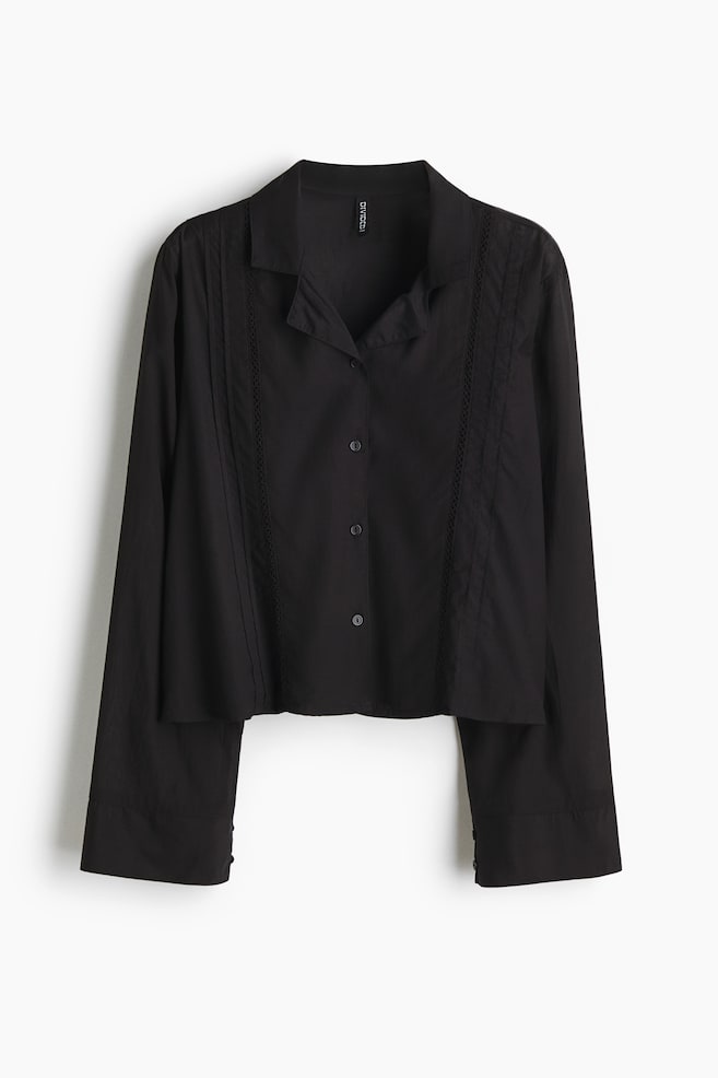 A black shirt is showcasing its FRONT, featuring a pointed collar, a button-up front with visible black buttons, and long sleeves with wide buttoned cuffs. Vertical pintucks are detailing the front panels, accompanied by lace trim, and the shirt is ending in a cropped, straight hem. - 1