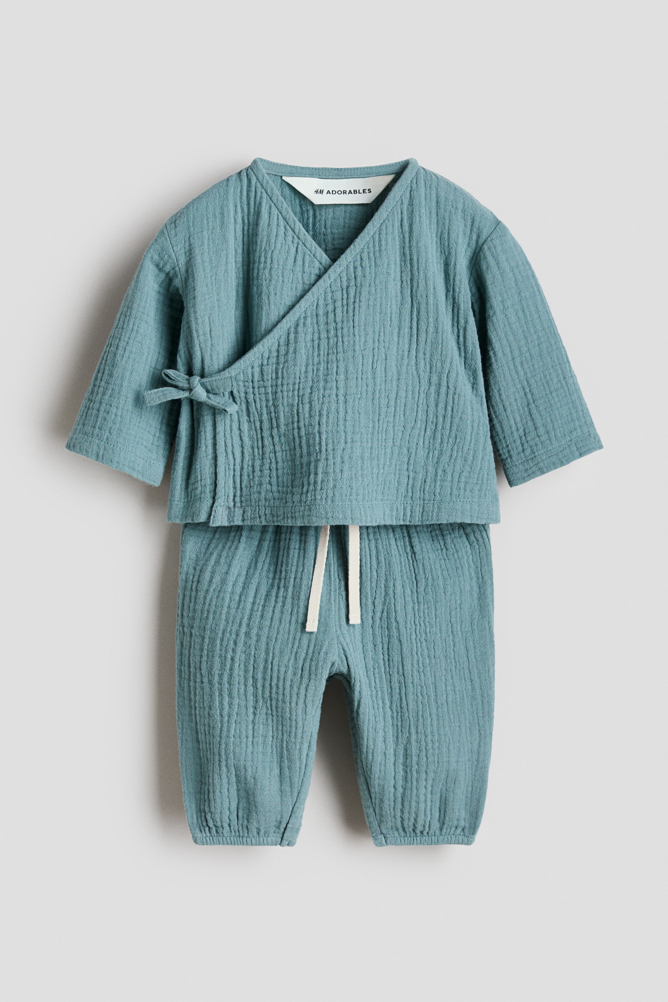 View larger image: A two-piece baby clothing set in a teal, crinkled fabric is displayed from the front. The top showcases a kimono-style wrap with a V-neck and a tie closure that forms a bow on the left side, while the matching pants feature an elasticated waistband with an off-white drawstring and elasticated cuffs at the ankles.