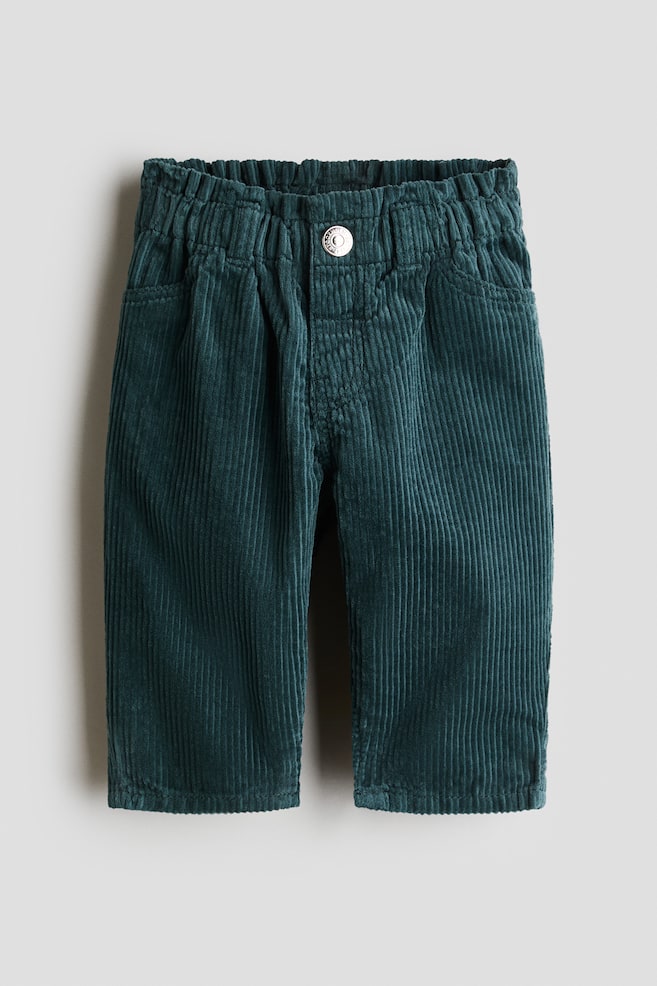 A pair of dark teal green corduroy pants is shown from the front, featuring an elasticated paperbag waist, a mock fly with a silver-tone button, and decorative front pockets. - 1