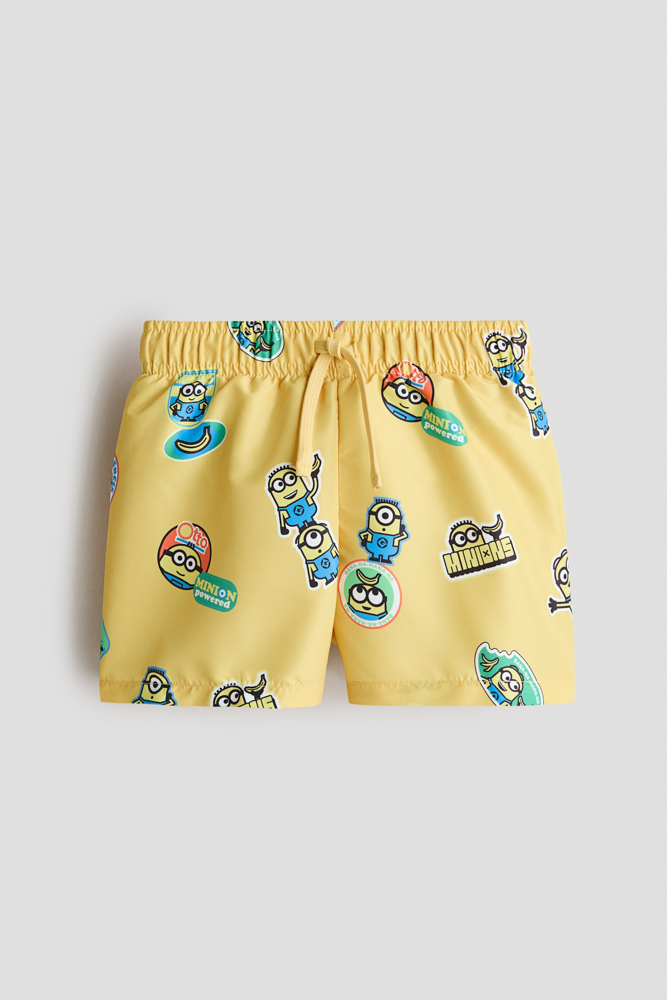 Größeres Bild ansehen: Badeshorts mit gelbem Grundton und einem Print-Motiv verschiedener Minions-Charaktere und Grafiken. Sie verfügen über einen verdeckten Gummizug und eine Kordel, die an der Vorderseite der Taille gebunden wird.