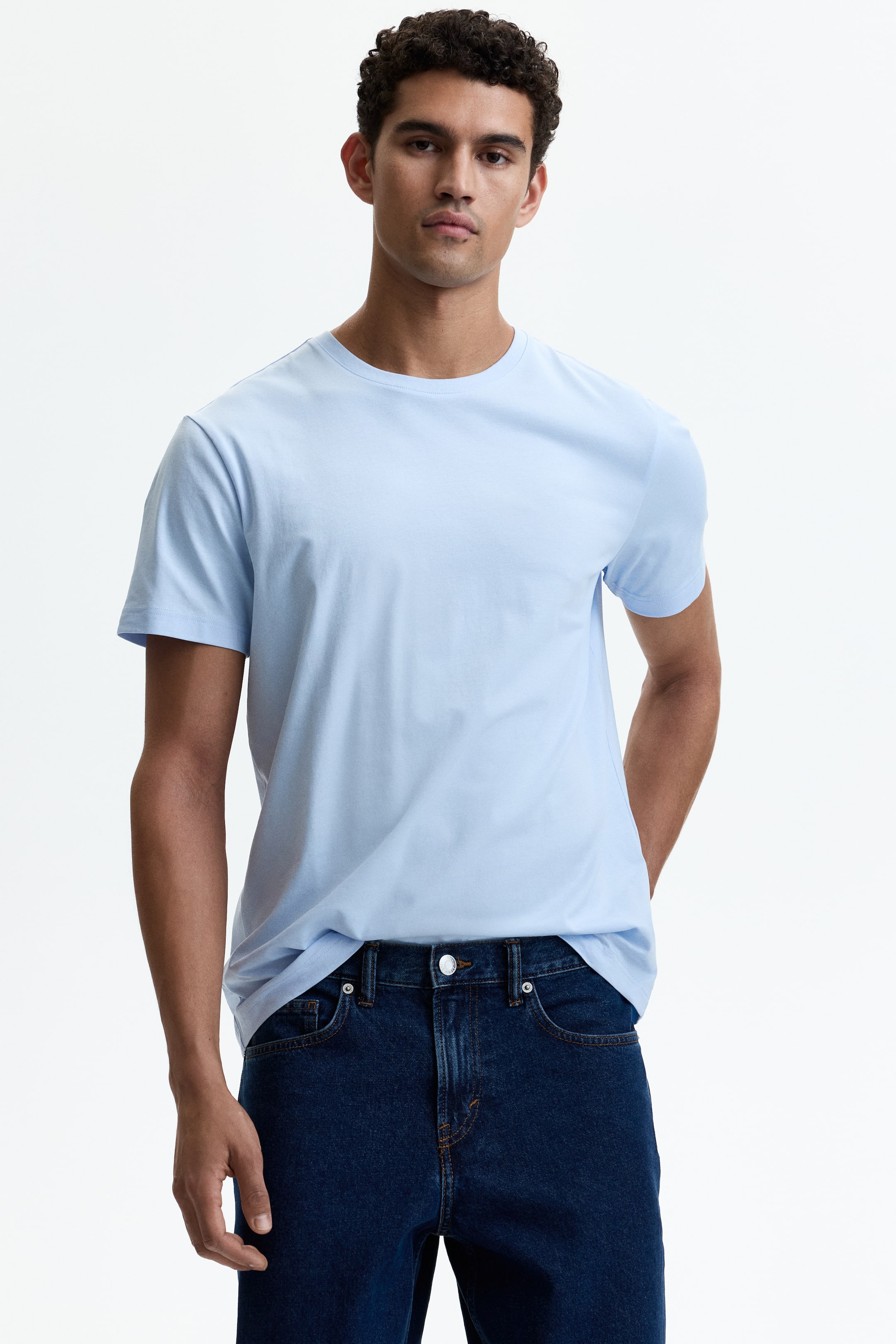 Größeres Bild ansehen: Ein hellblaues, figurbetontes T-Shirt mit einem runden, gerippten Halsausschnitt und einem gerade geschnittenen Saum wird vom Model getragen und ist in dunkelblaue Jeans gesteckt.