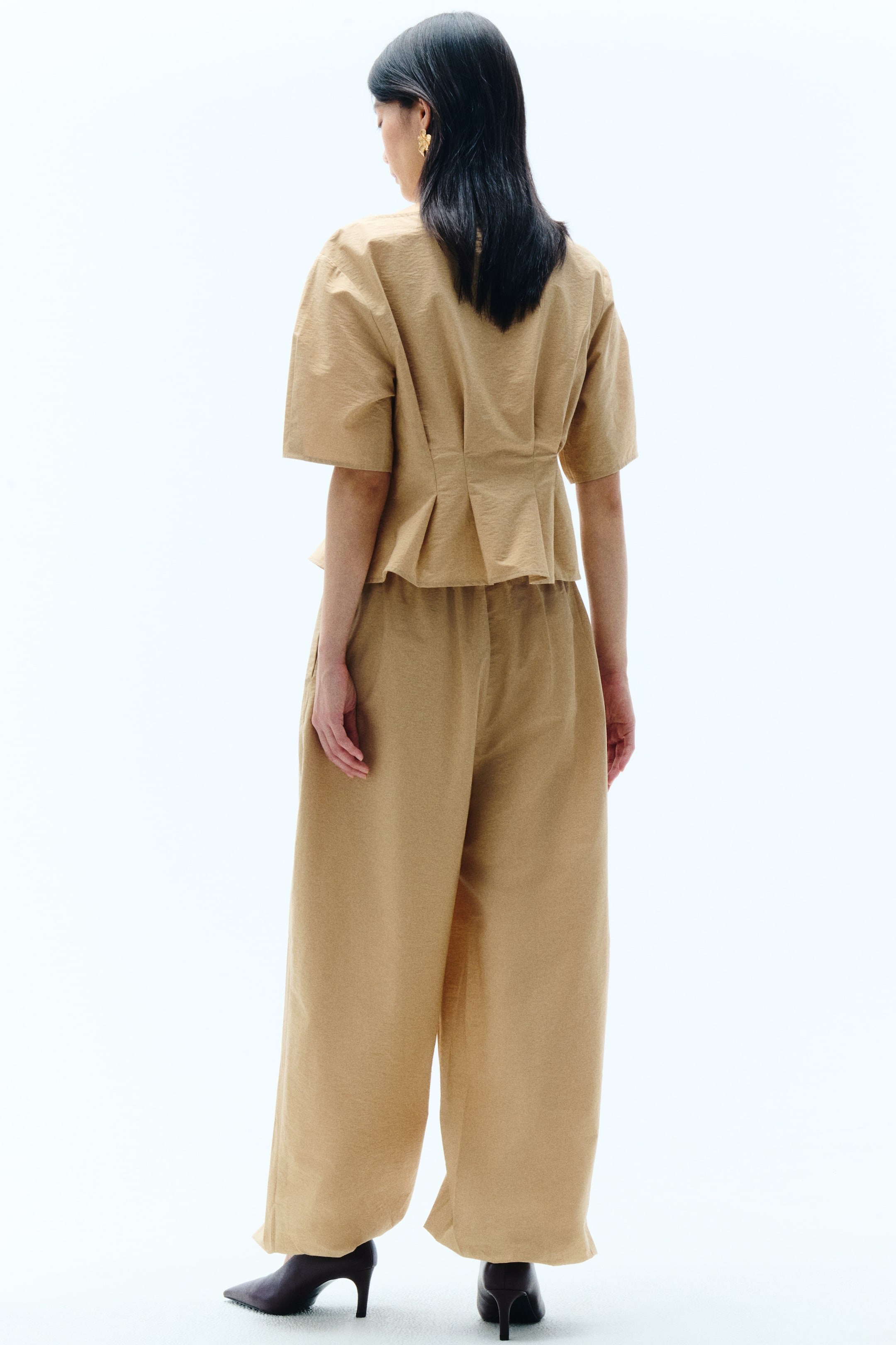 View larger image: A tan, textured two-piece set is shown from the back. The short-sleeved top features a fitted waist with pleats that create a flared peplum. The matching wide-leg trousers come with an elasticated waistband and a flowy silhouette.