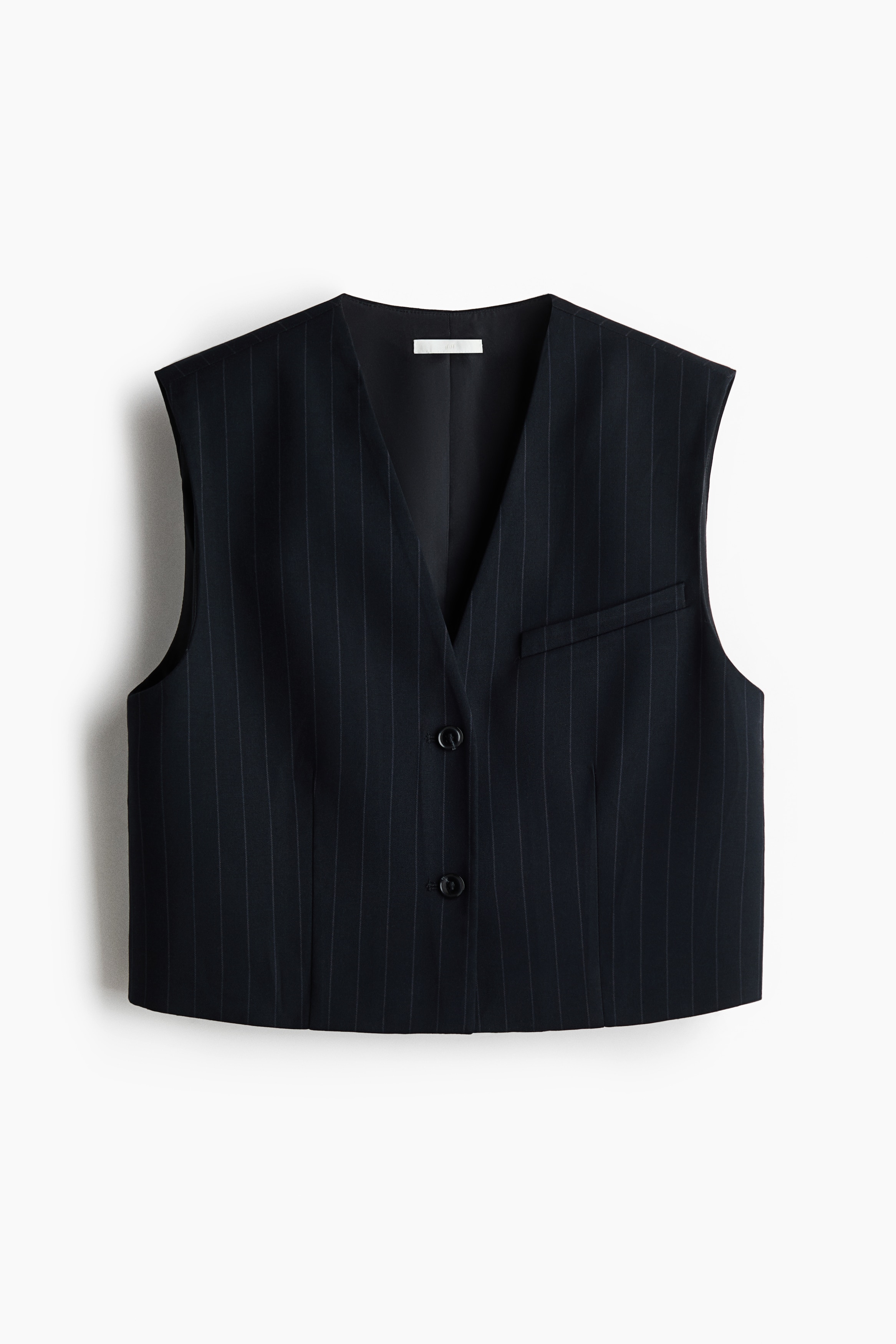 A cropped black pinstripe vest is being worn by the model, featuring a deep V-shaped neckline, structured shoulders, and a front button closure. The sleeveless vest has a single welt chest pocket and is paired with a matching pinstripe mini skirt.