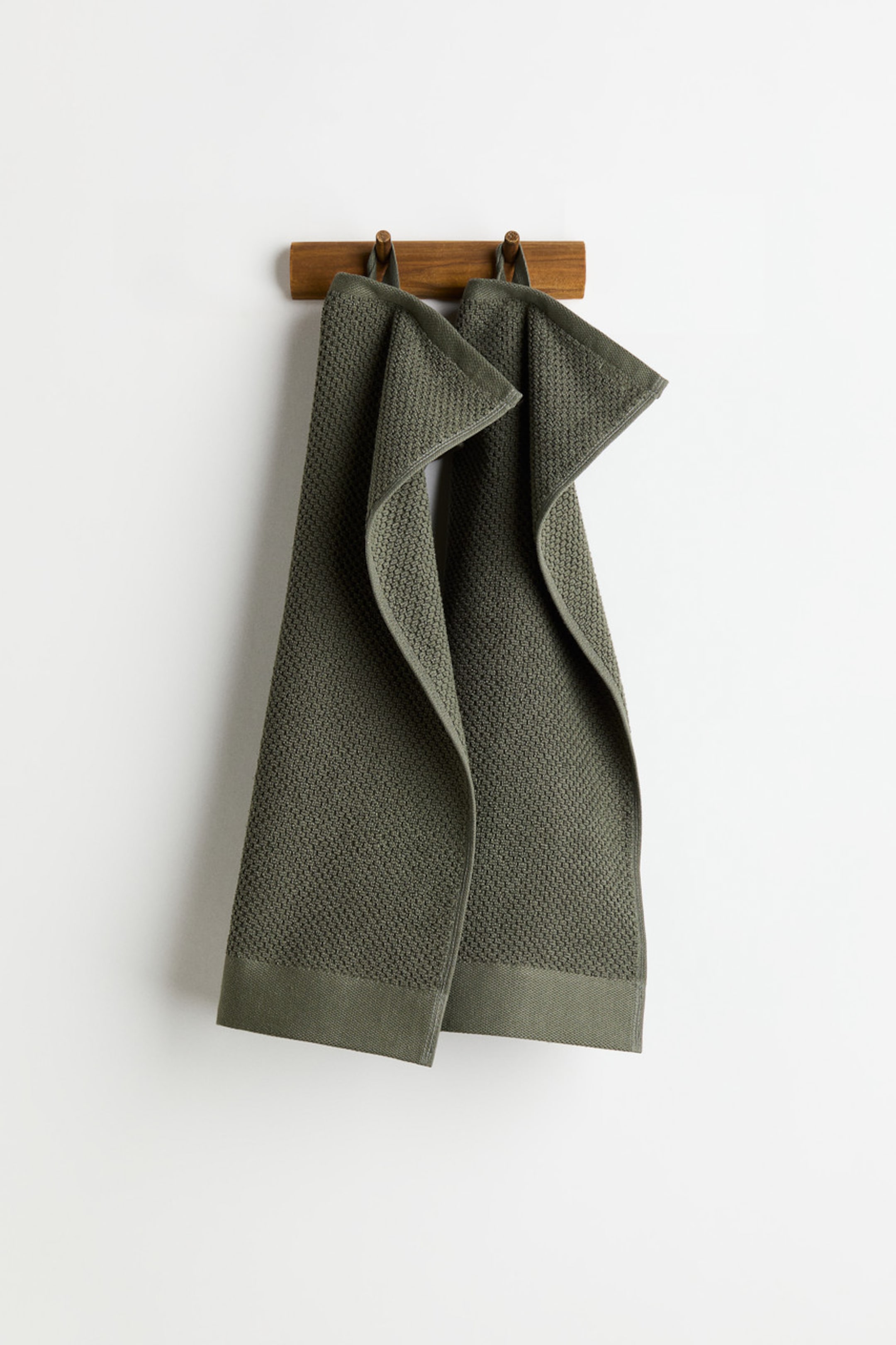 View larger image: Two olive green waffle-weave towels are hanging, folded in half lengthwise, from a wooden rack. Each towel is displaying a smooth, solid band along its bottom edge.
