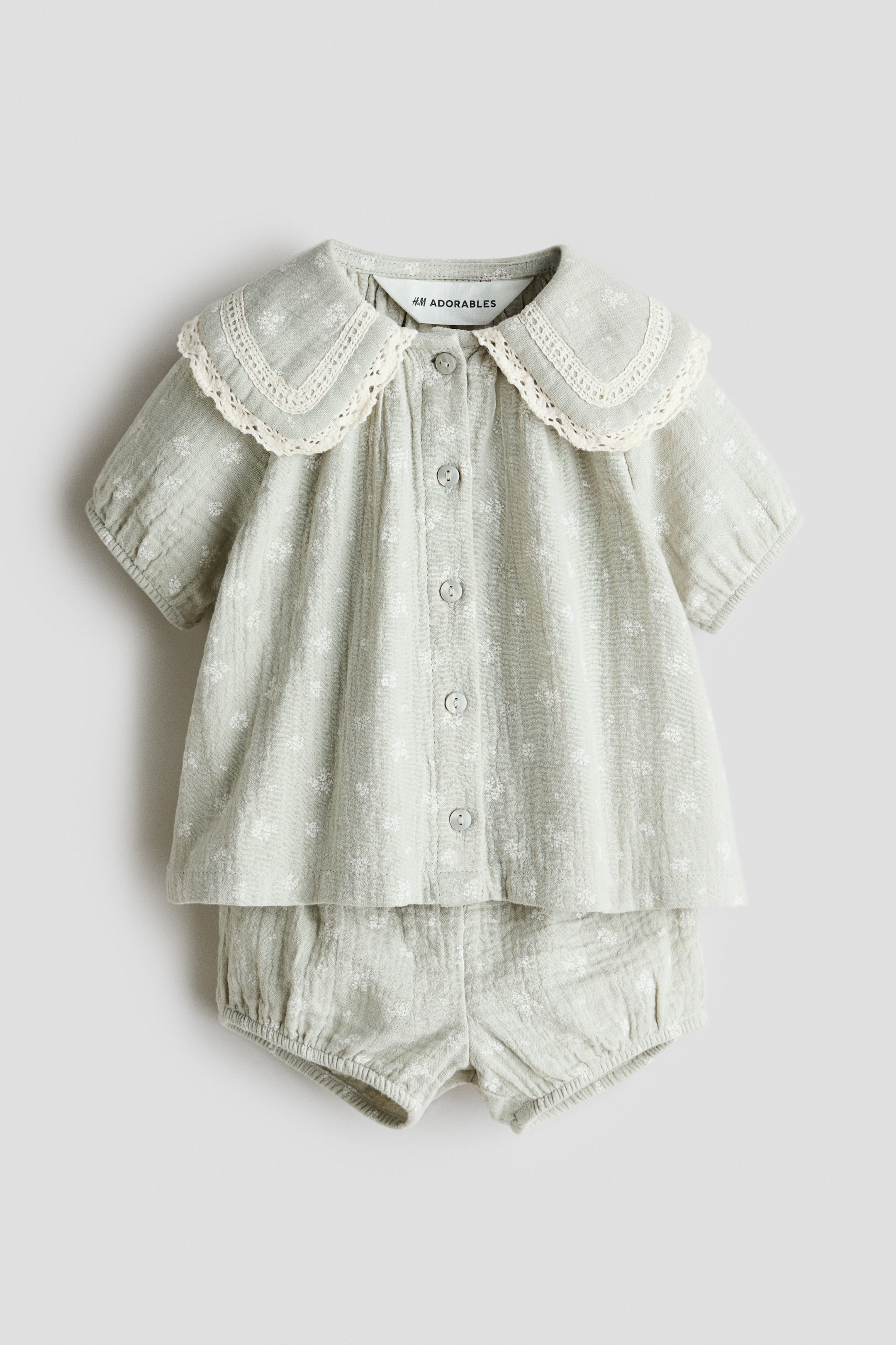 View larger image: A baby outfit featuring a light green top with matching bloomer shorts, both printed with a small white floral pattern. The top features a wide scalloped collar trimmed with lace, short puffed sleeves and a front button fastening, while the shorts have elasticated leg openings.
