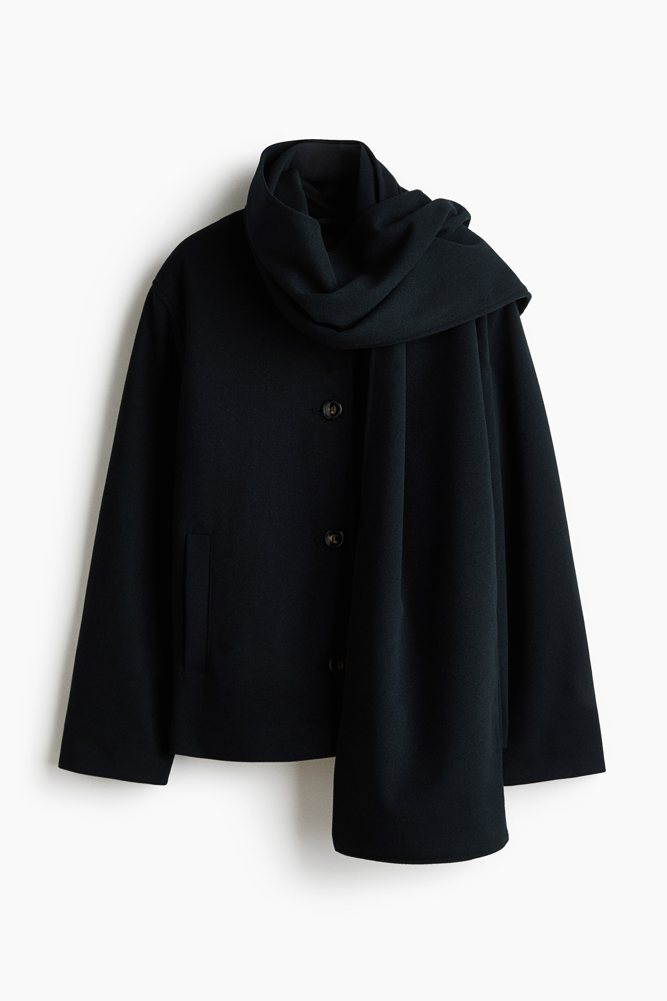 Visa större bild: En kort, svart oversized jacka hänger framtill och är tillverkad i ett mjukt, filtat tyg, med knappknäppning fram, nedhasade axlar och en vertikal passpoalficka i sidan. En matchande lång scarf är virad runt halsringningen och faller ned framtill över den raka fållen.