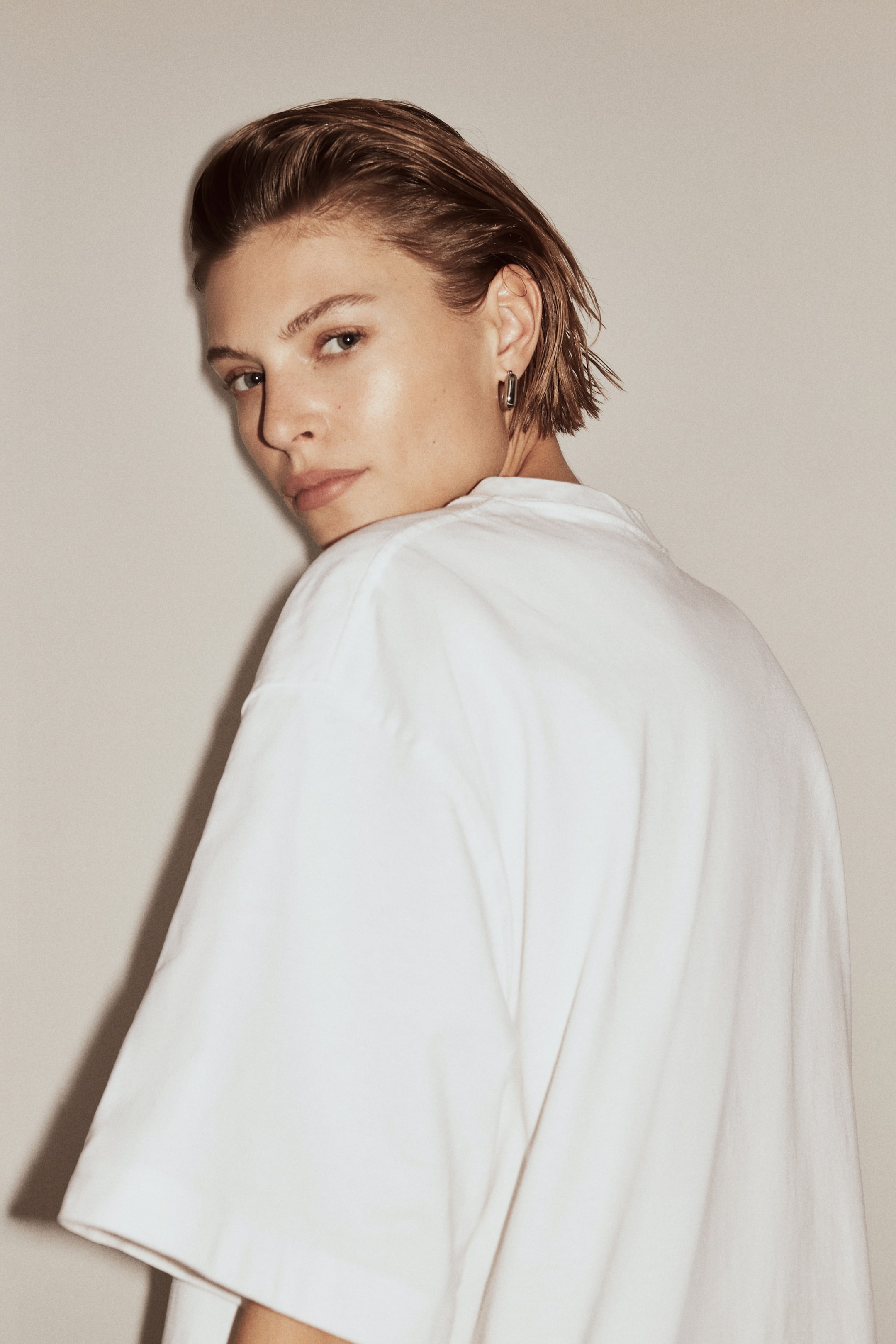 拡大画像を表示: 白のオーバーサイズTシャツを着用しており、リラックスしたフィット感と幅広のショートスリーブが際立っています。長方形のシルバー調フープイヤリングが耳たぶを飾っています。