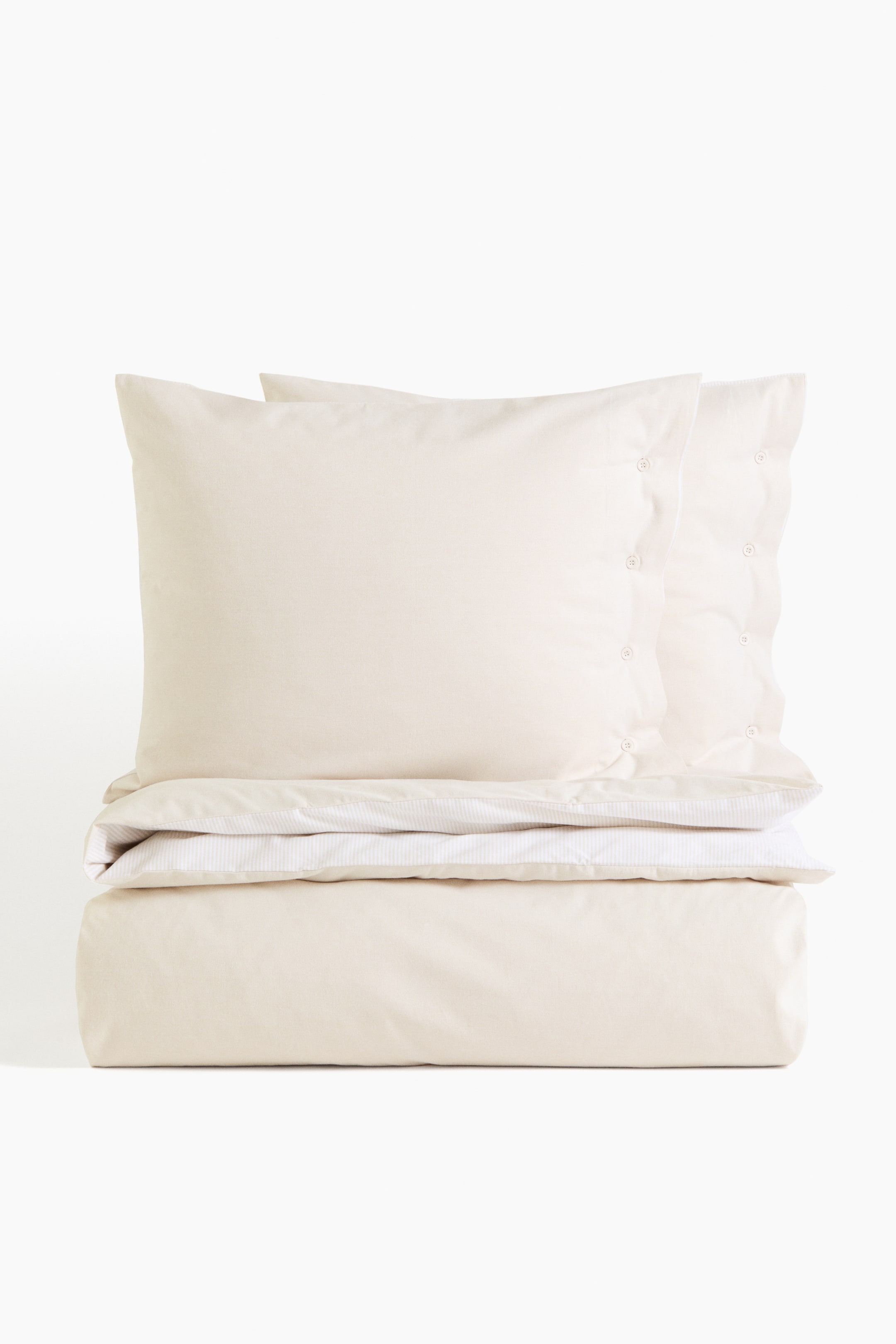 View larger image: A light cream bedding set features two pillowcases with visible button closures and a neatly folded duvet cover, revealing a subtle striped lining along its top edge.