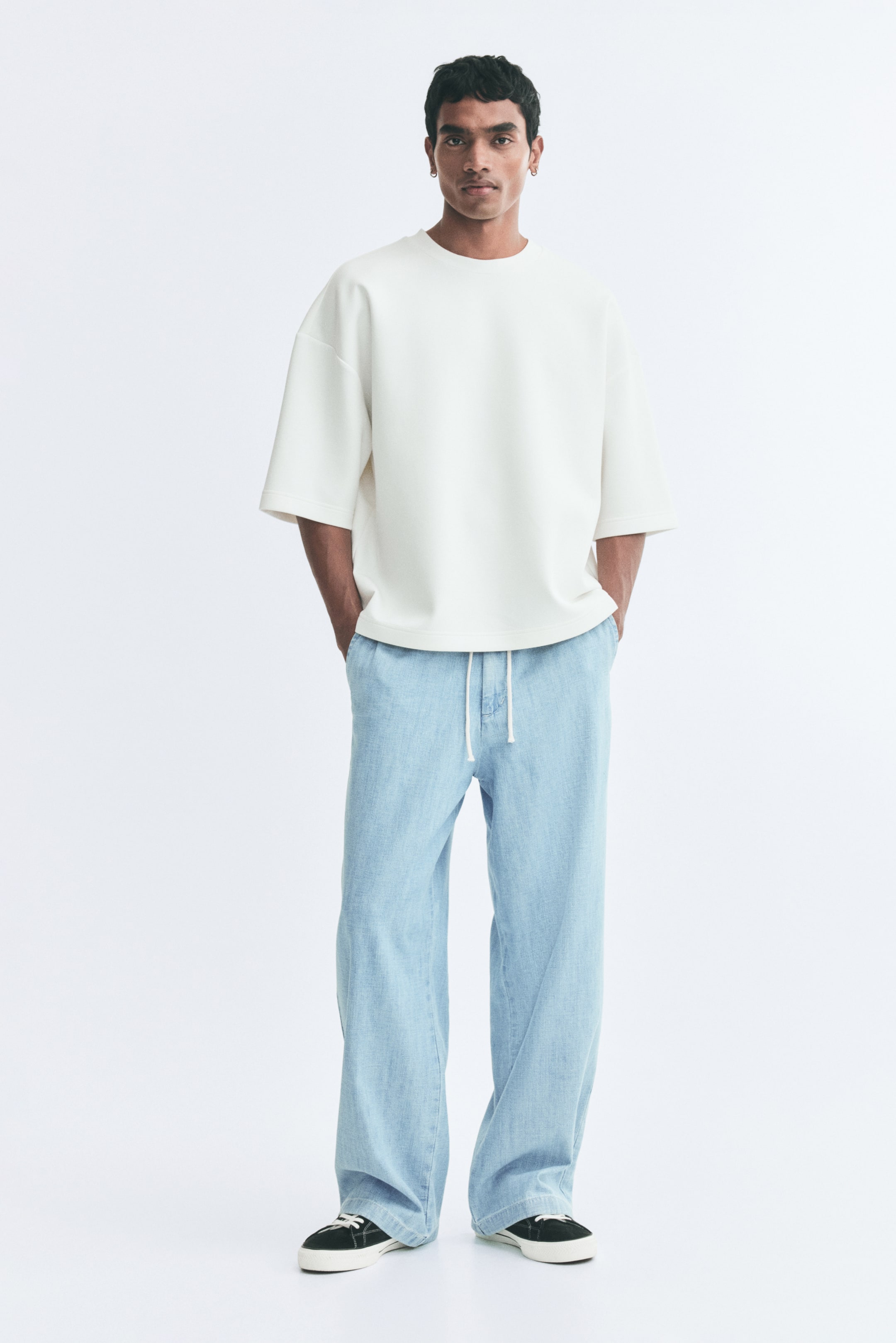 View larger image: The model is wearing light blue denim pants with a relaxed fit, an elasticated waistband with a white drawstring, and diagonal side pockets. The pants are styled with an off-white, short-sleeved T-shirt and black sneakers, while the model stands with hands tucked into the pockets.