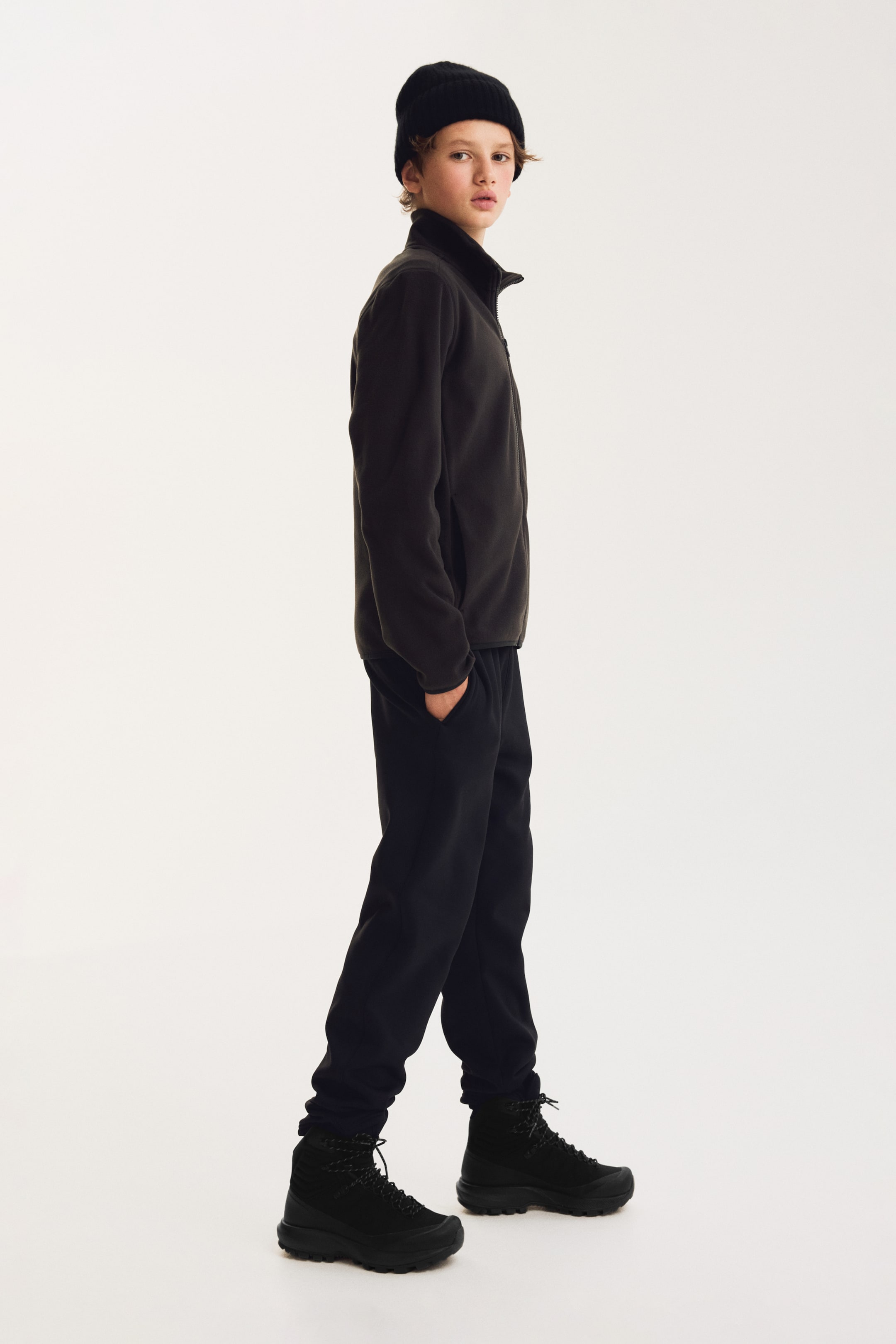 View larger image: A dark grey fleece jacket featuring a high stand-up collar and a full-length zip is shown in side profile. The jacket is paired with matching black jogger trousers featuring elasticated cuffs that sit above black lace-up boots.