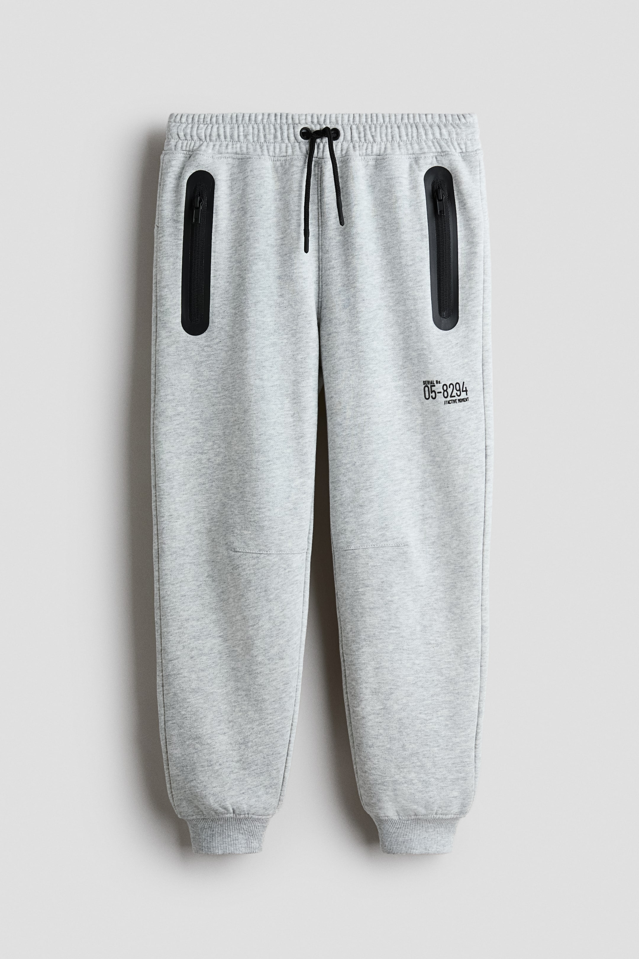 View larger image: Light grey sweatpants are shown from the front, featuring an elastic waistband with a black drawstring and two black vertical zippered pockets on the upper front. The pants also display horizontal seams across the knees, ribbed cuffs at the ankles, and a black text graphic on the right thigh.