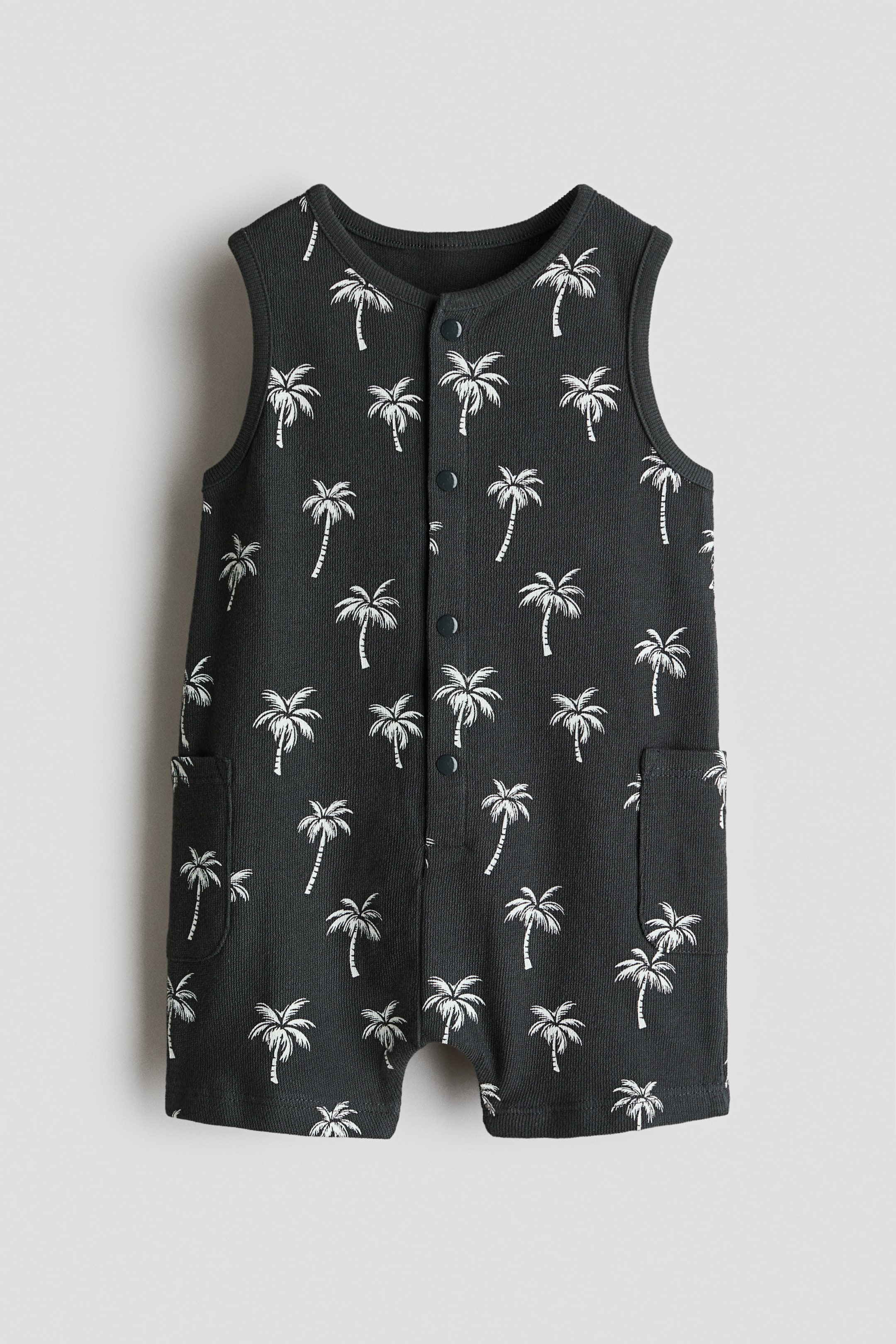 View larger image: A sleeveless romper suit is showcasing its front, featuring a dark grey fabric adorned with white palm trees. It is crafted with a round neckline, press-studs running down the front, and two patch pockets on the sides, finishing with short legs.