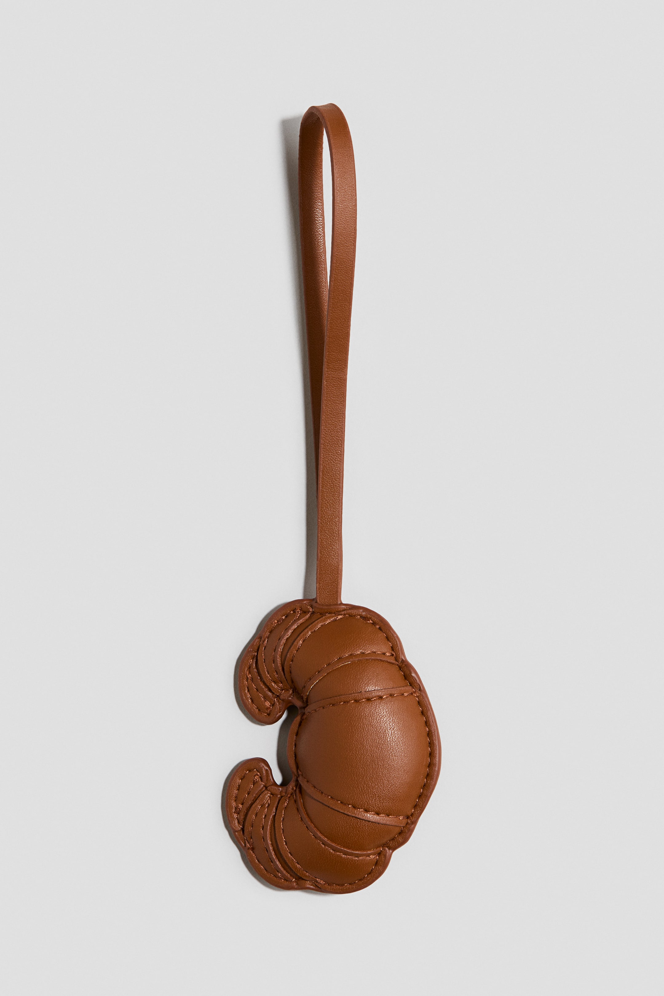 Voir une image plus grande: Une breloque matelassée pour sac, en forme de croissant, présente sa face avant. Elle est confectionnée en tissu enduit brun, avec des sections bien délimitées par des coutures, et une fine boucle se prolonge à son sommet pour la fixation.