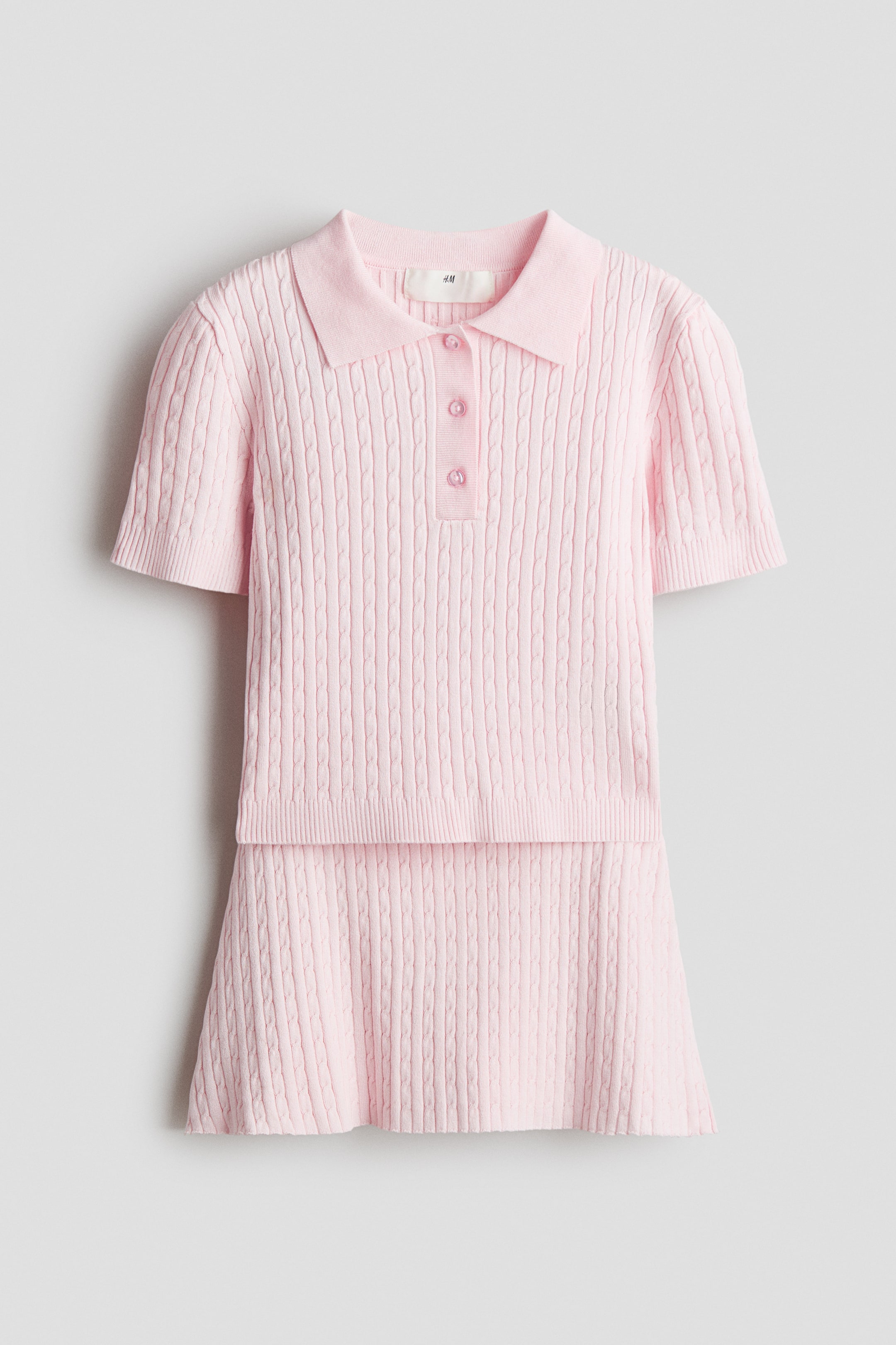 Agrandir l'image: Un ensemble en maille torsadée rose est présenté de face, comprenant un polo à manches courtes avec col, patte de boutonnage à trois boutons, et bords-côtes aux poignets et à l'ourlet, ainsi qu'une jupe assortie avec taille élastiquée et un léger évasement à l'ourlet.