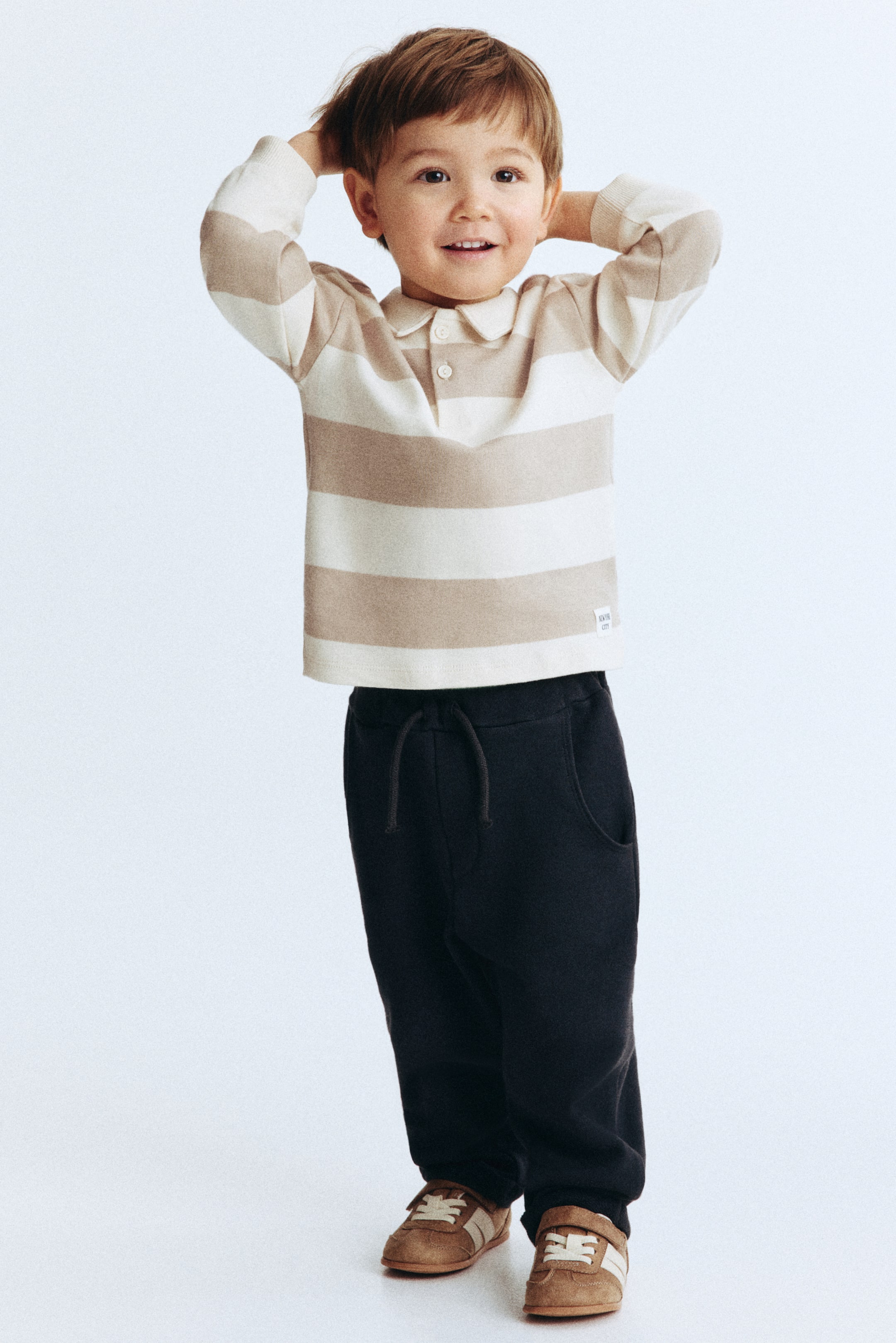 Visa större bild: En långärmad pikétröja med alternerande horisontella ränder i beige och kräm bärs framifrån och är matchad med mörkgråa sweatpants. Pikétröjan har en spetsig krage, en knappslå med två knappar och ribbade muddar, medan sweatpantsen är utformade med sidofickor och en synlig dragsko i midjan.