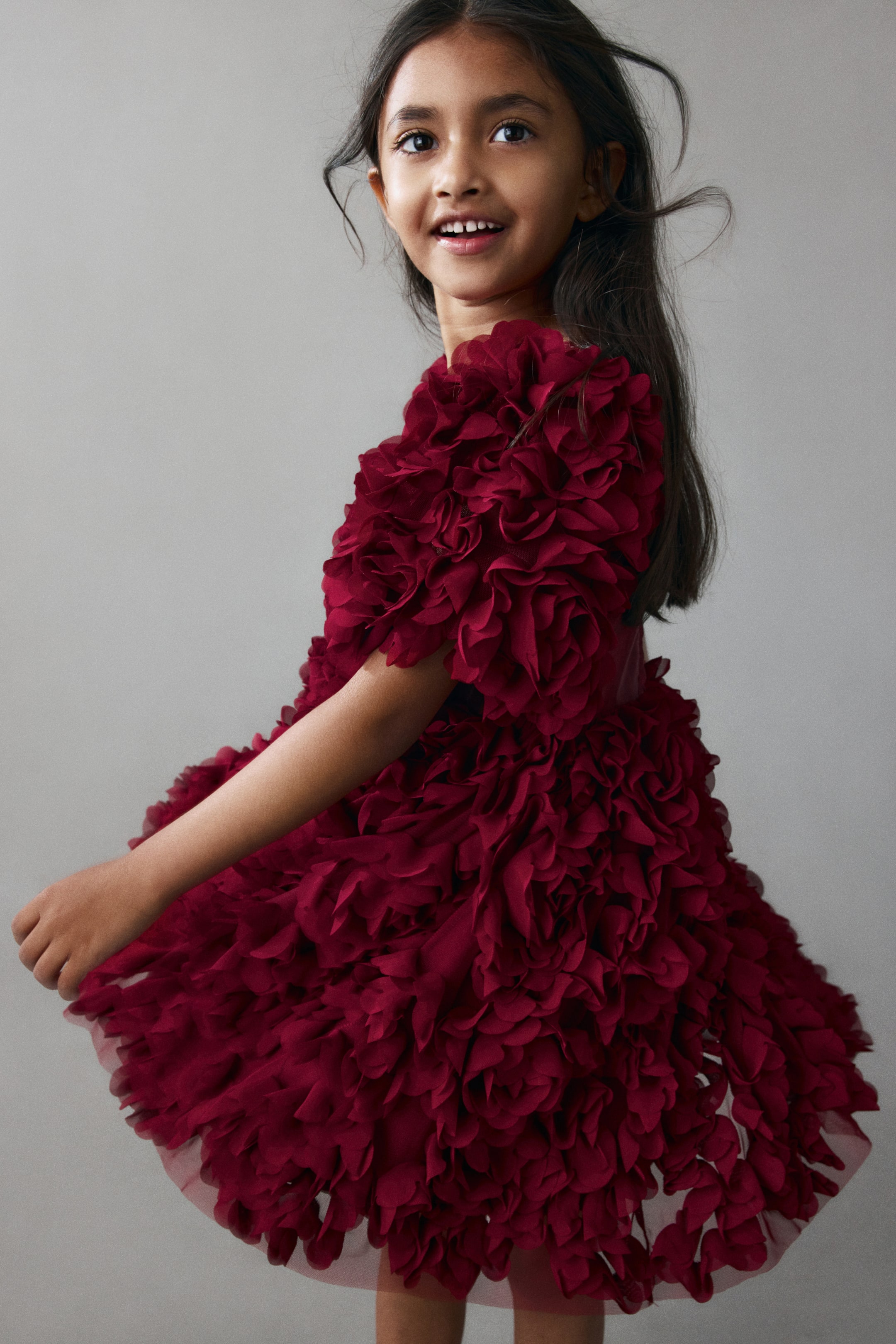 View larger image: A dark red dress is shown from a side angle, featuring dense, three-dimensional fabric ruffles that cover the entire garment from the short sleeves to the flared skirt. The dress presents a highly textured, voluminous design while being worn.