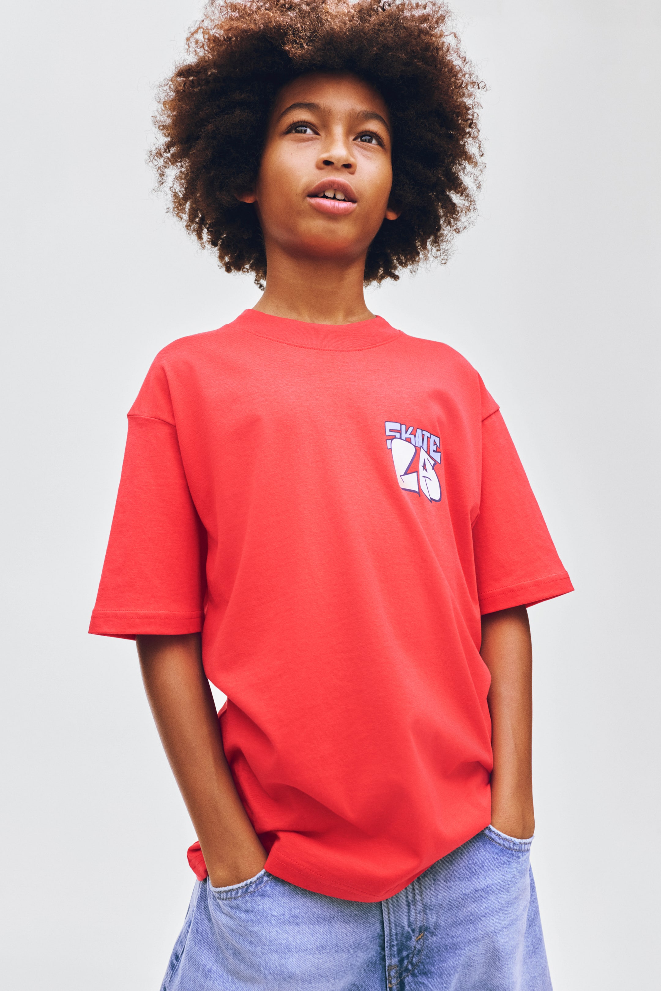 View larger image: The red t-shirt showcases a crew neck and relaxed short sleeves. It sports a graphic design with 'SKATE 25' in white and blue on the left chest, draping casually over light blue denim jeans.