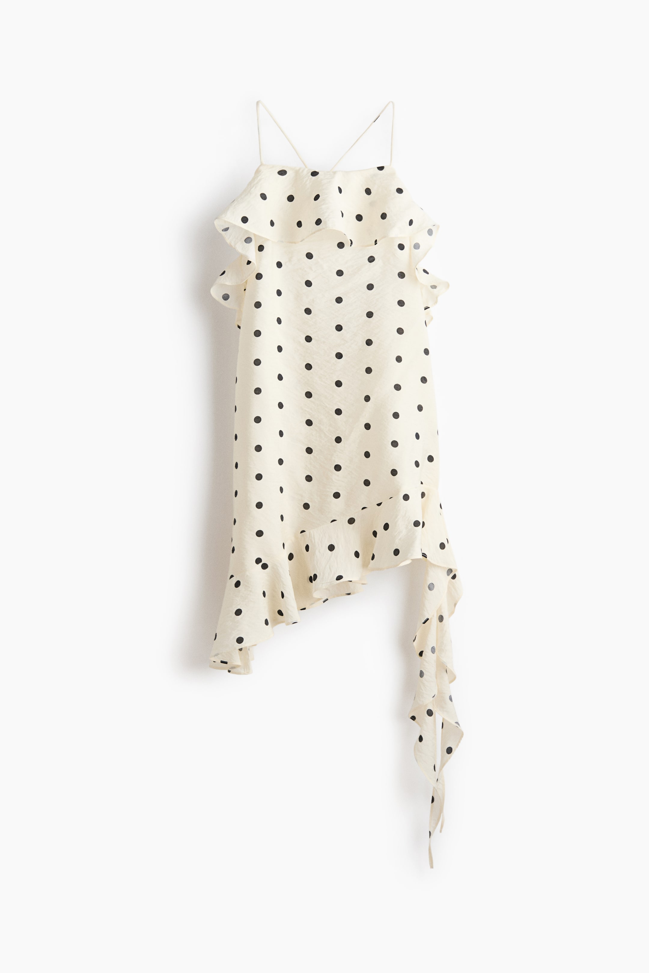 Ampliar la imagen: Una blusa de tirantes con estampado de lunares en color crema está mostrando su FRENTE. Presenta finos tirantes de espagueti y un volante que se extiende por el pecho y los hombros. Un dobladillo asimétrico exhibe un volante en cascada en el lado derecho.