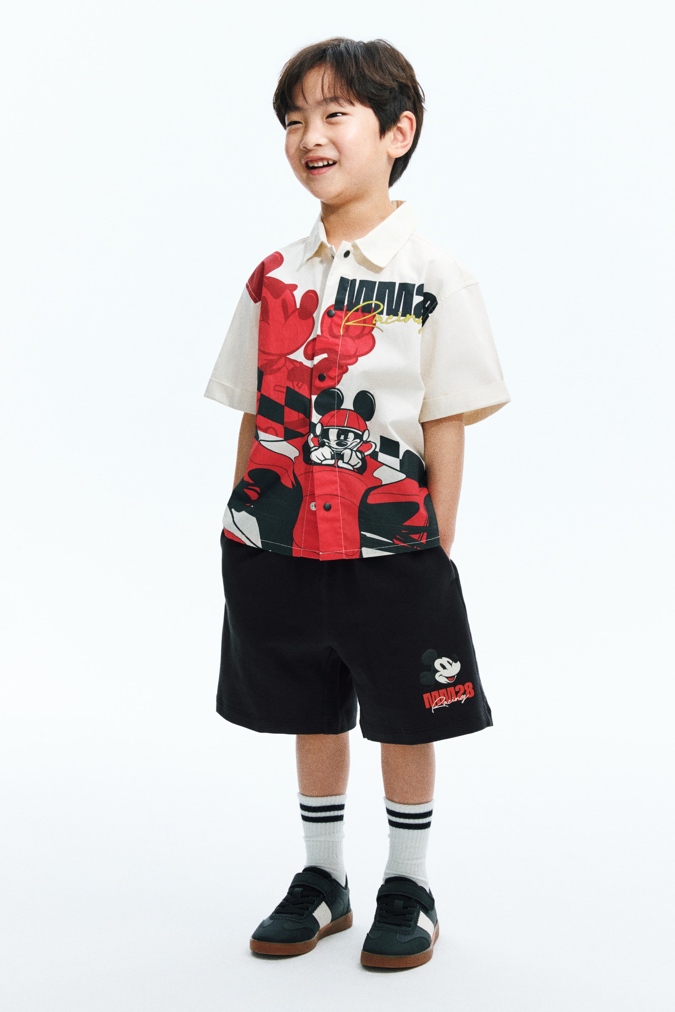 View larger image: A short-sleeved button-up shirt in off-white, adorned with a red and black checkered Mickey Mouse racing graphic on the front, is paired with black shorts featuring a small Mickey Mouse and 'Racing' design on the left leg. Completing this child's ensemble are white crew socks with black stripes and black sneakers with white side stripes and velcro closures, all styled to face slightly to the right.