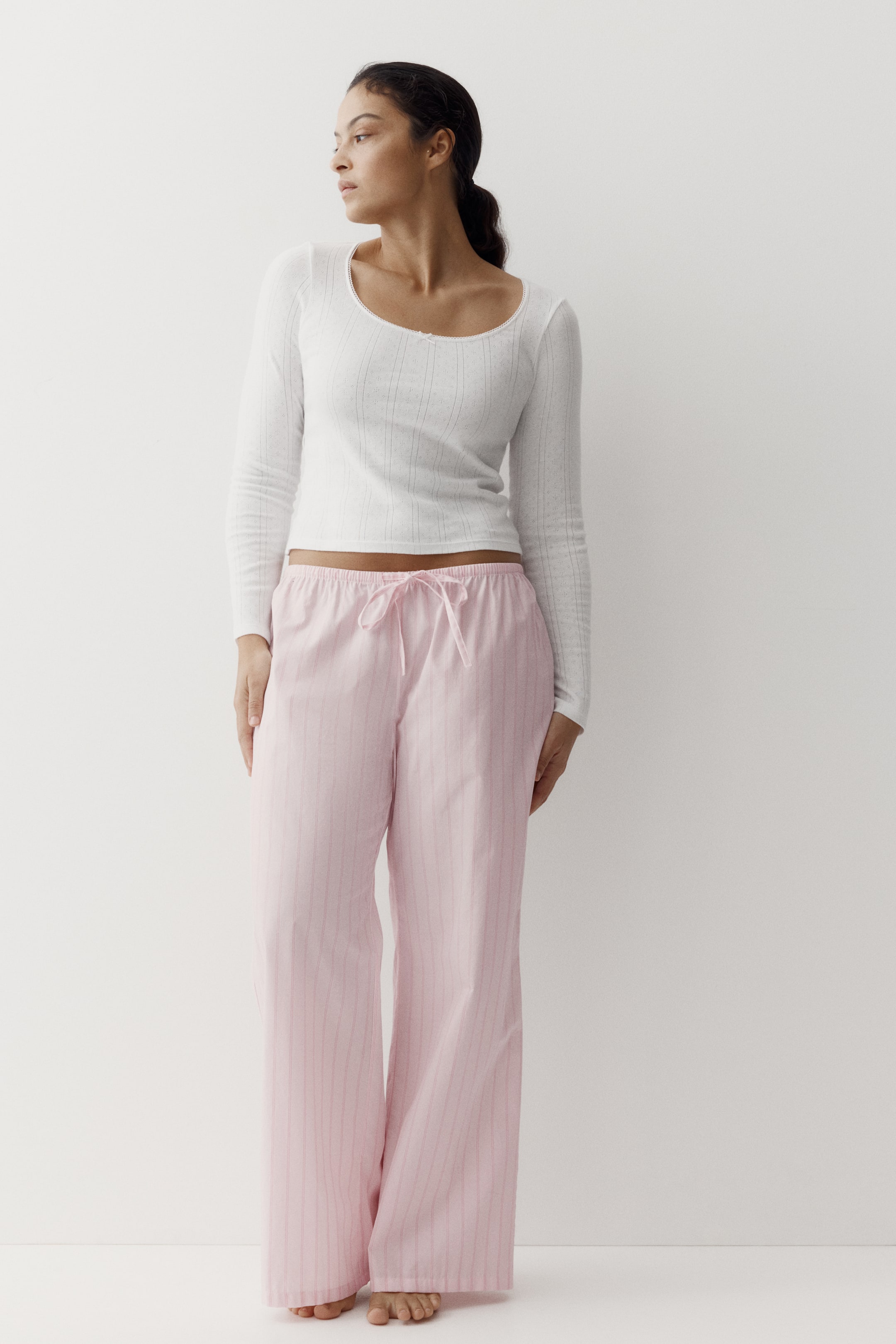 Ver imagen más grande: Una camiseta blanca de manga larga con cuello redondo acanalado, que es corta, se lleva con pantalones de pijama rosa de pierna ancha con sutiles rayas verticales y cintura con cordón atada en un lazo.