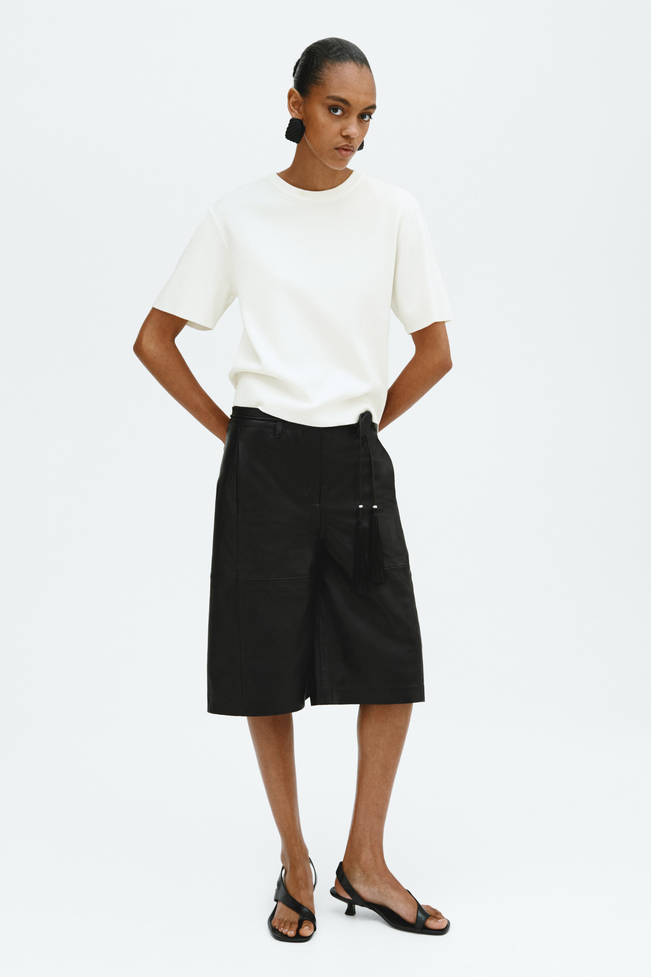 Grotere afbeelding bekijken: Een wit T-shirt met ronde hals en korte mouwen wordt gedragen, ingestopt in zwarte knielange culottes. De culottes hebben zijzakken, stikdetails en een decoratieve zwarte kwast met twee witte kralen die aan de linkerheup hangen.