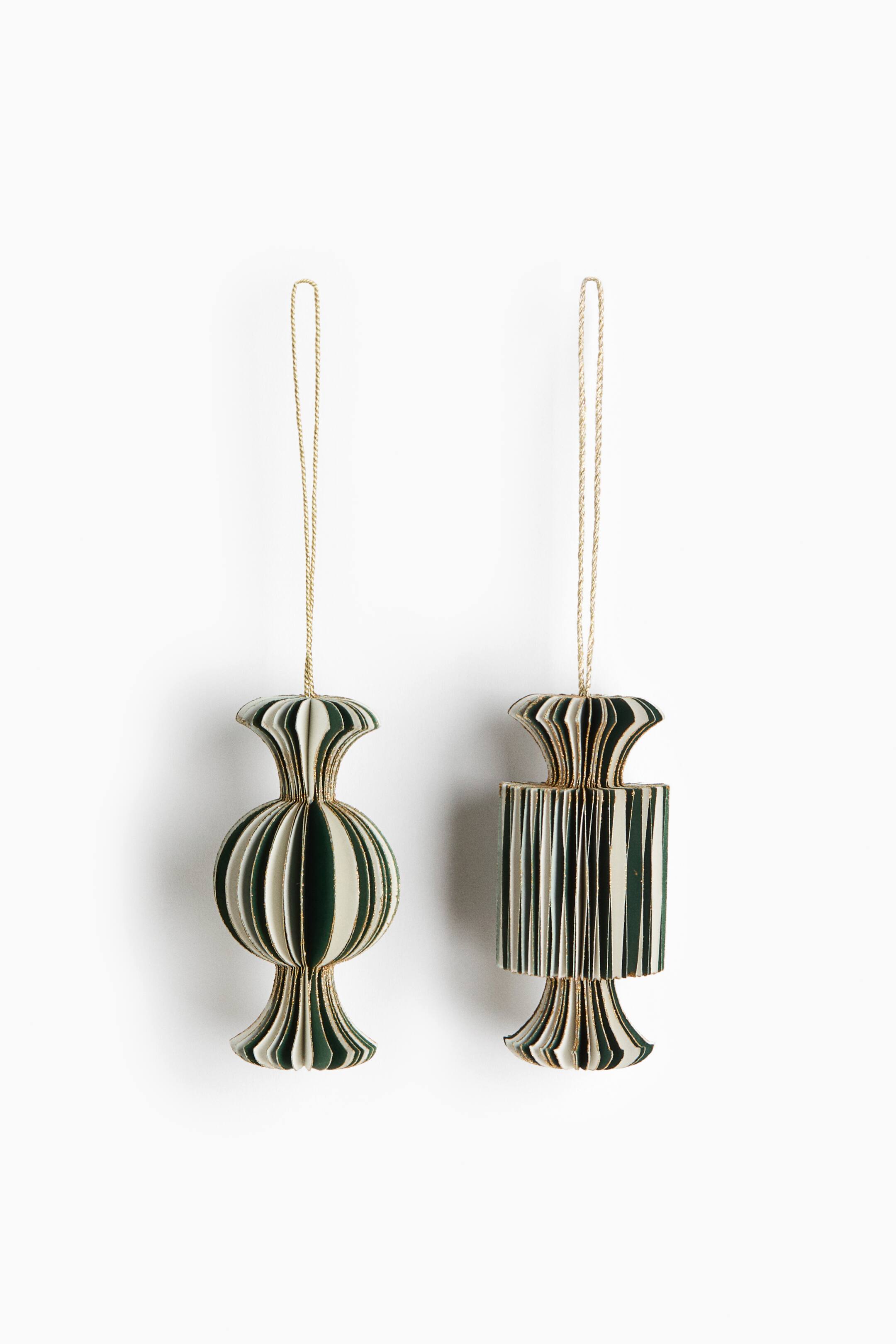 View larger image: Two pleated paper ornaments hang side by side, showing a front view of their accordion-style designs with alternating dark green and cream stripes and gold-glittered edges. The left ornament features a bulbous, spherical center, while the right ornament showcases a cylindrical center, with both designs tapering into cinched, flared ends suspended by thin gold strings.