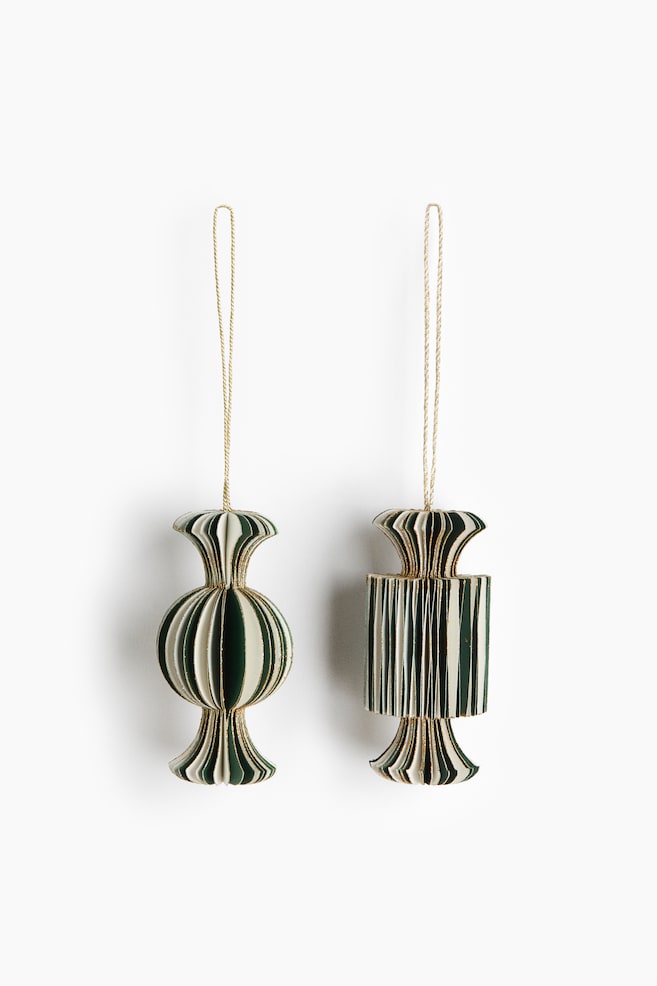Two pleated paper ornaments are hanging side-by-side, displaying a front view of their accordion-style designs with alternating dark green and cream stripes and gold-glittered edges. The left ornament is featuring a bulbous, spherical center, while the right ornament is showcasing a cylindrical center, with both designs tapering into cinched, flared ends suspended by thin gold strings. - 1