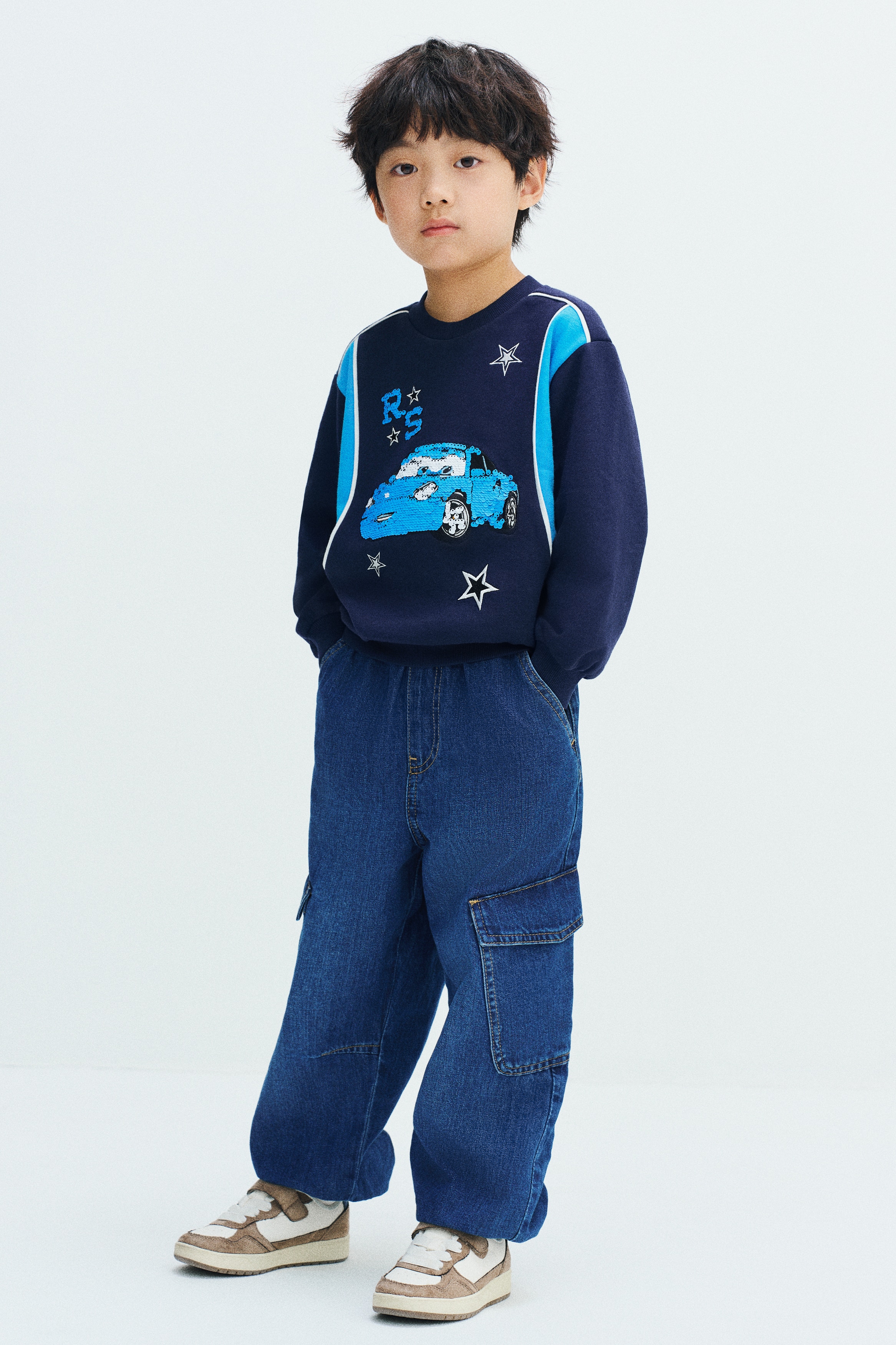 Denim joggers in a washed blue cotton fabric are shown from the front, featuring a gathered elasticated waistband, a faux fly, and diagonal side pockets. The balloon-style legs feature flap-covered cargo patch pockets on the sides and taper down to gathered elasticated hems.