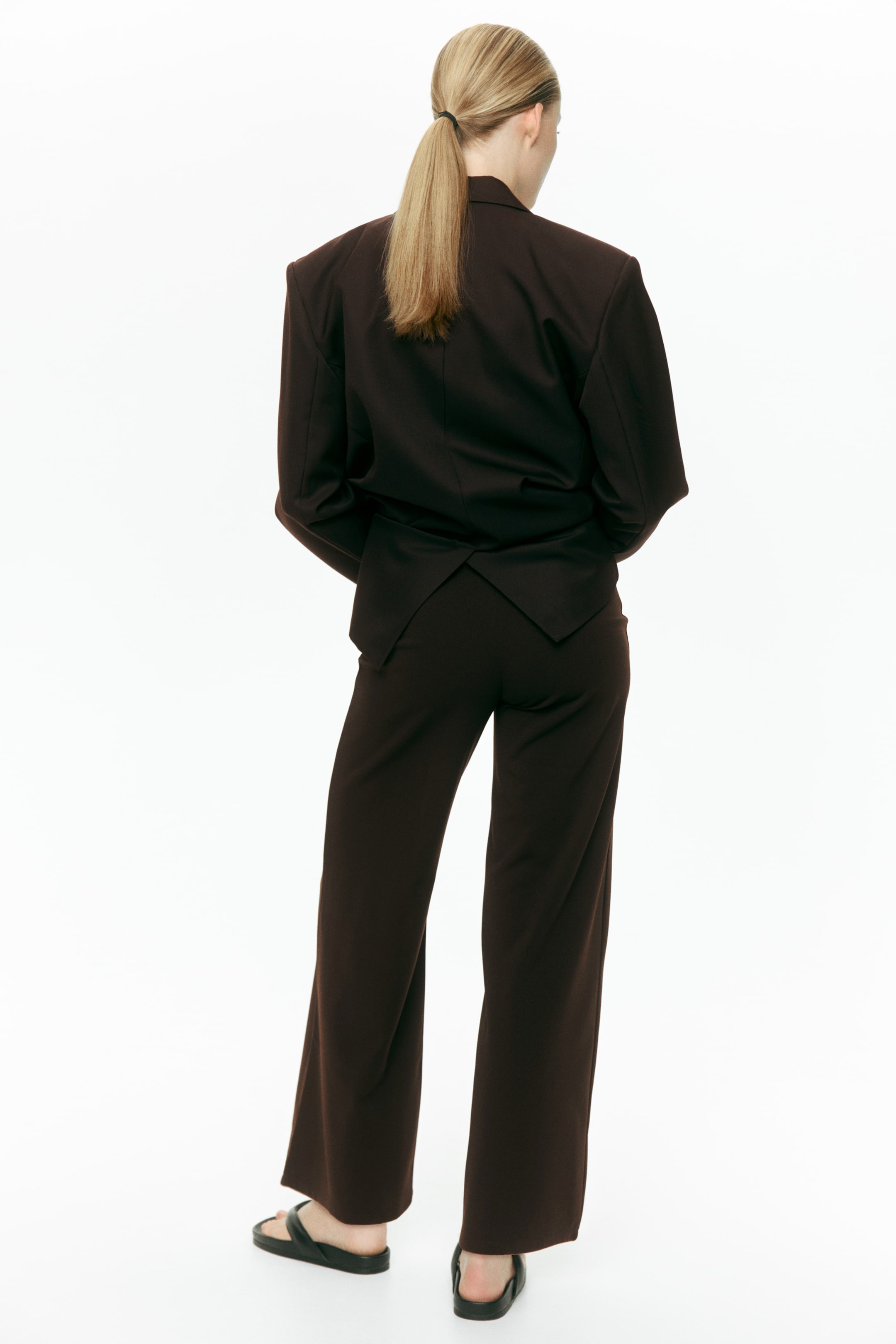 Agrandir l'image: Un blazer marron foncé et un pantalon large assorti sont présentés de dos, le blazer arborant des épaules structurées et un ourlet arrière en forme de V. Le pantalon long affiche une coupe large, porté avec des sandales plates noires.