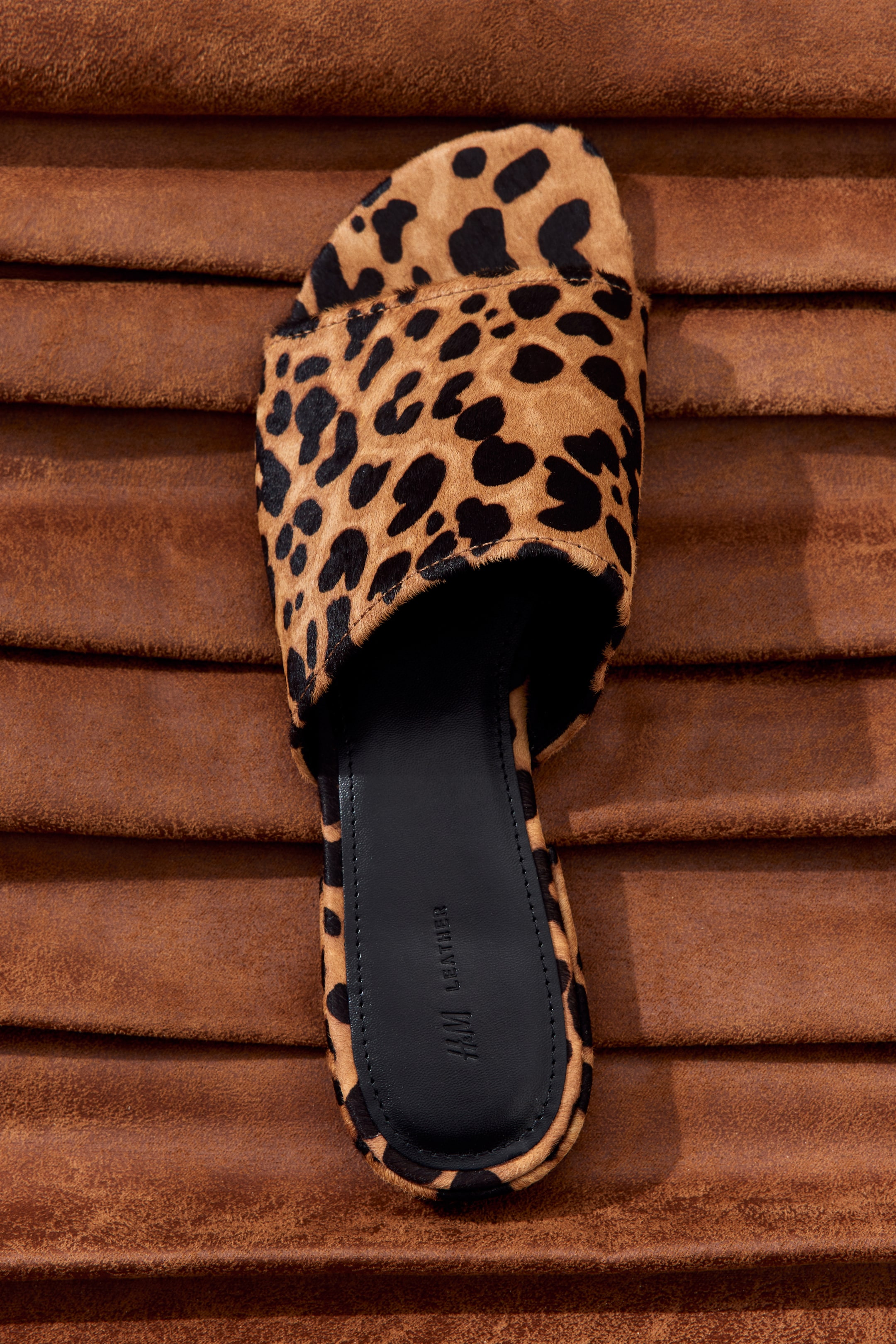 Visa större bild: En leopardmönstrad slidesandal presenteras på en texturerad brun yta. Sandalen visar upp en bred rem med svarta leopardfläckar på en brun bas, och dess svarta fotbädd är präglad med "H&M Leather"-märkning.