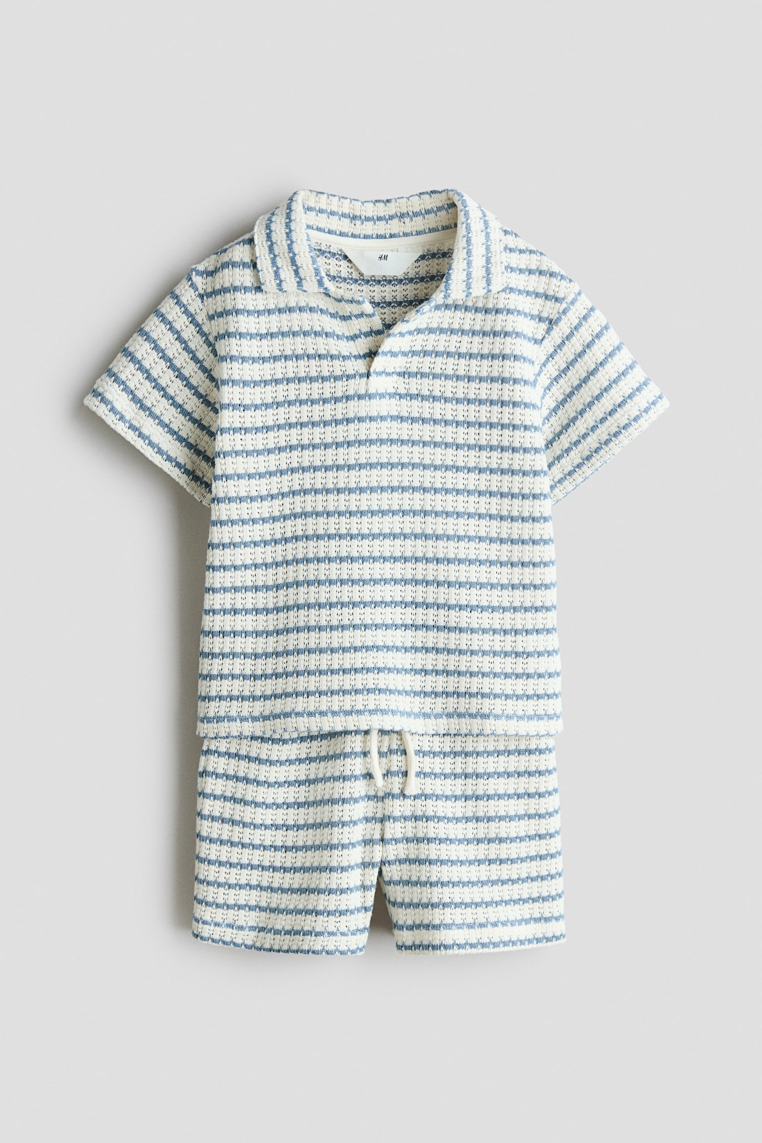 A two-piece knit set is facing front, featuring a short-sleeved polo shirt and matching shorts in a textured, blue and cream horizontal stripe pattern. The shirt is displaying a collar with an open, buttonless placket, and the shorts are featuring an elastic waistband with a white drawstring tied at the front.