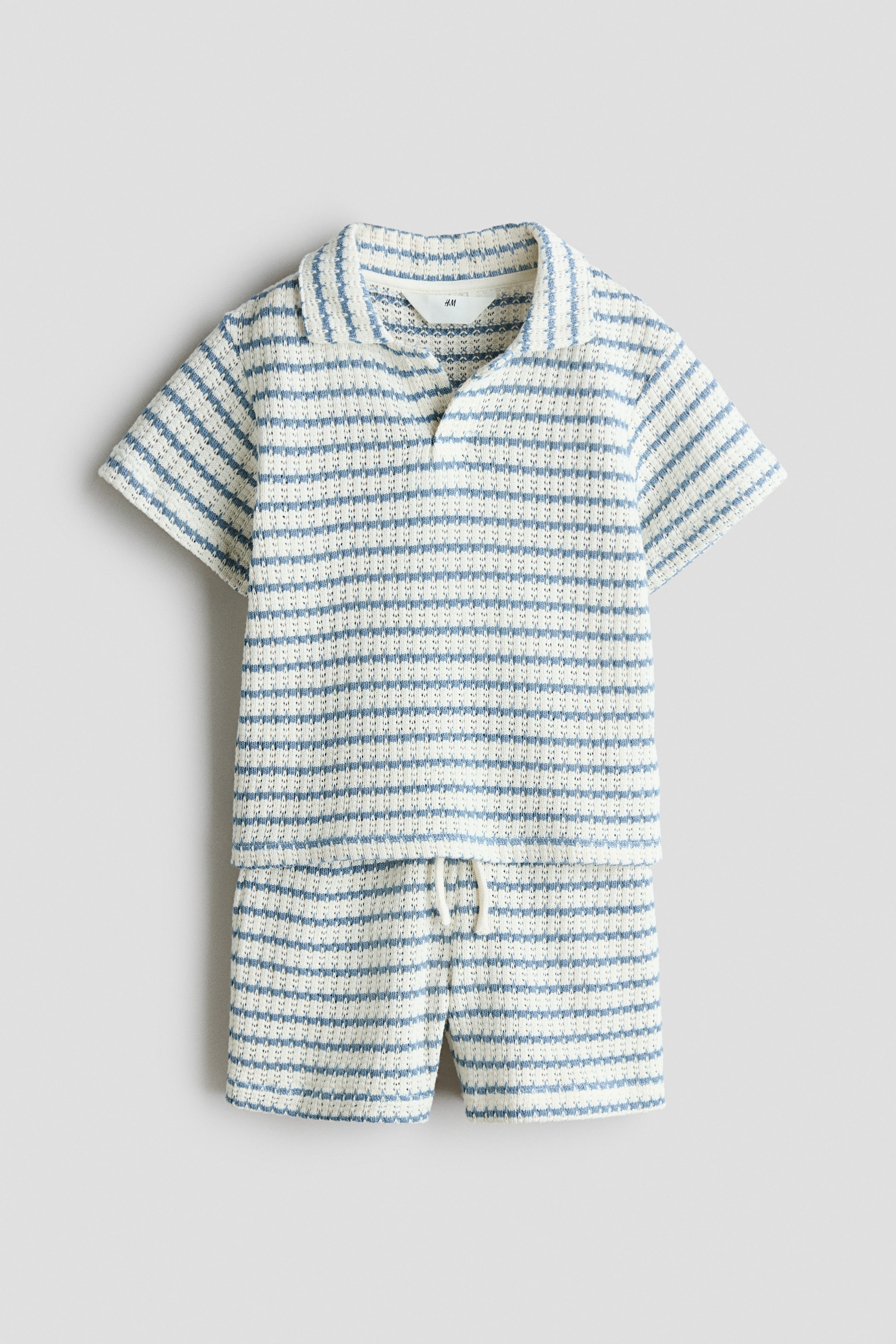 View larger image: A two-piece knit set is shown from the front, featuring a short-sleeved polo shirt and matching shorts in a textured blue and cream horizontal stripe pattern. The shirt features a collar with an open, buttonless placket, and the shorts feature an elastic waistband with a white drawstring tied at the front.