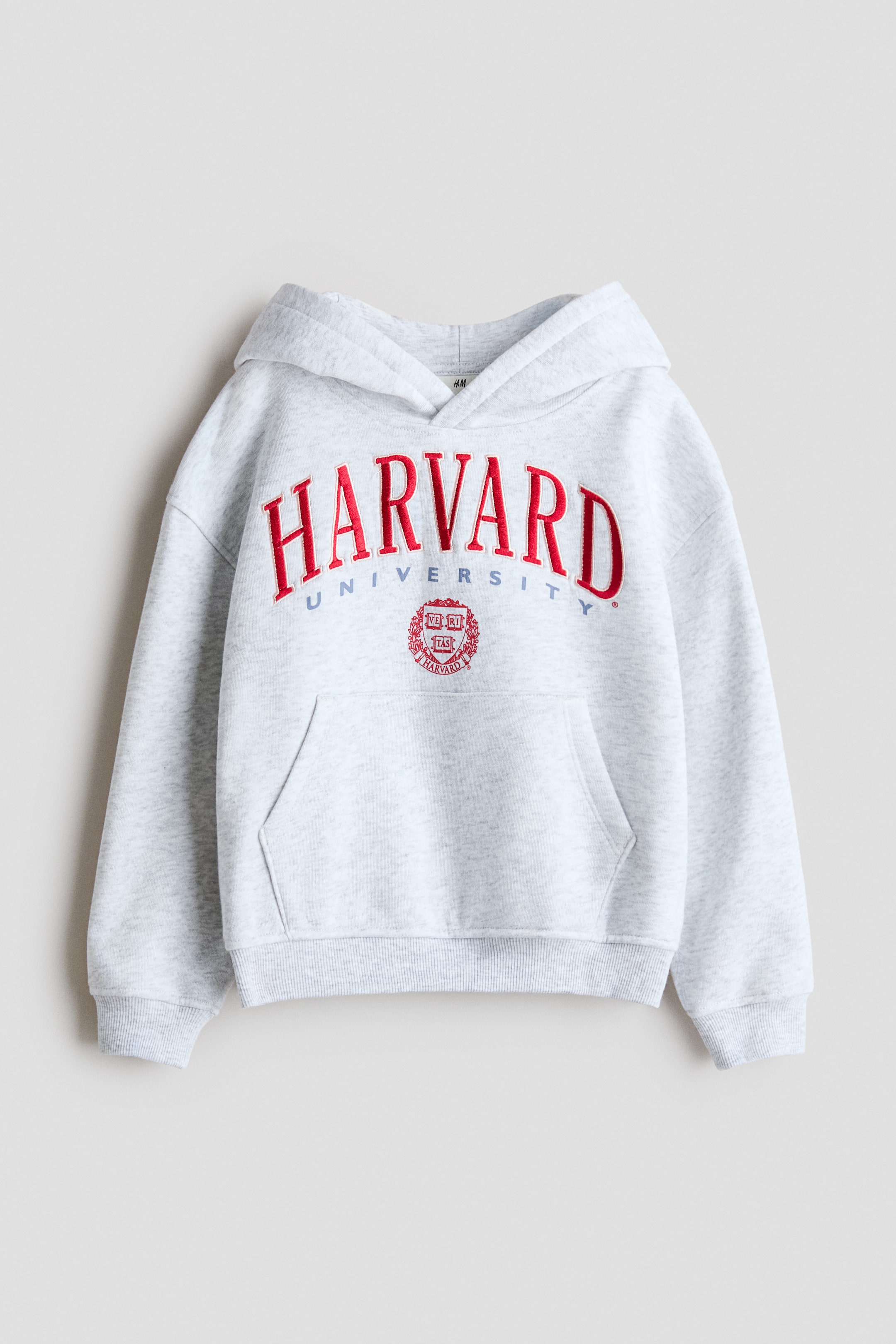 Agrandir l'image: Un sweat à capuche chiné gris clair est présenté de face, avec « HARVARD » en grandes lettres brodées rouges, « UNIVERSITY » en lettres bleues plus petites et un écusson circulaire brodé rouge en dessous. Il est doté d'une capuche, de manches longues, d'une poche kangourou, ainsi que de poignets et d'un ourlet côtelés.