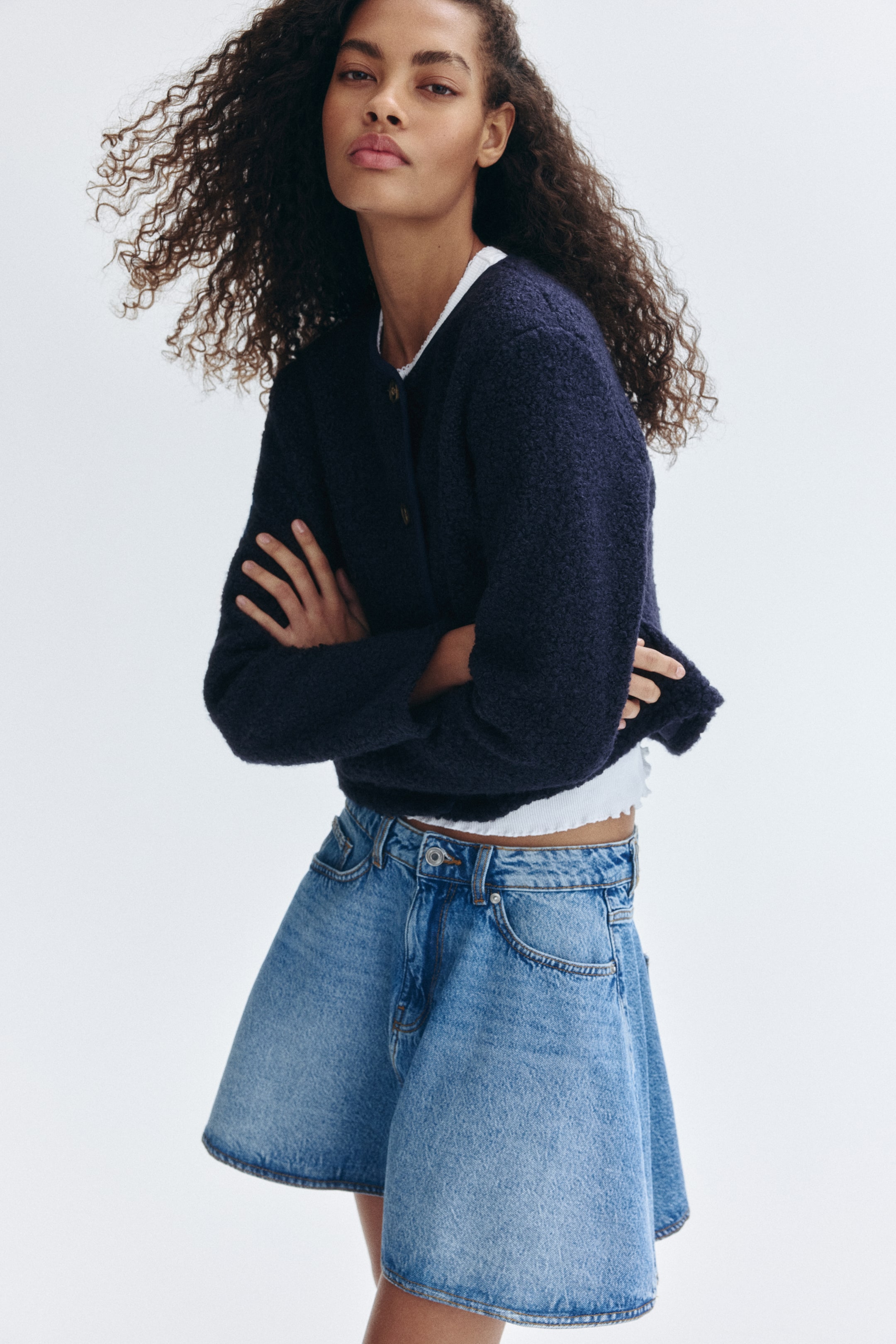 View larger image: A navy blue textured knit cardigan with a round neckline and long sleeves is being worn by the model. The cropped cardigan, featuring front buttons, is layered over a white top, and the model is crossing their arms.