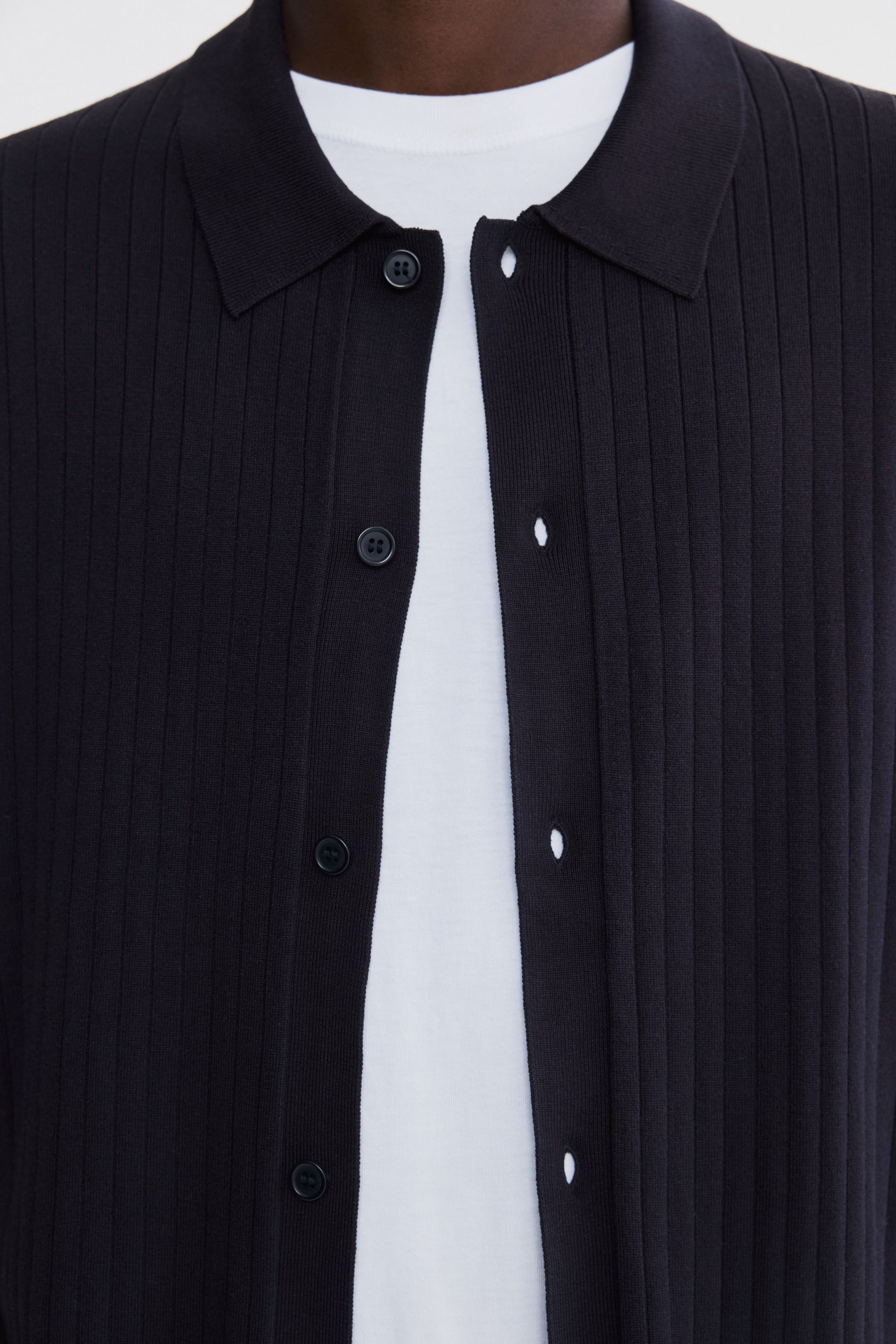 Agrandir l'image: Un cardigan en maille côtelée noire est porté ouvert sur un t-shirt blanc, affichant un col pointu et une patte de boutonnage complète avec des boutons noirs.