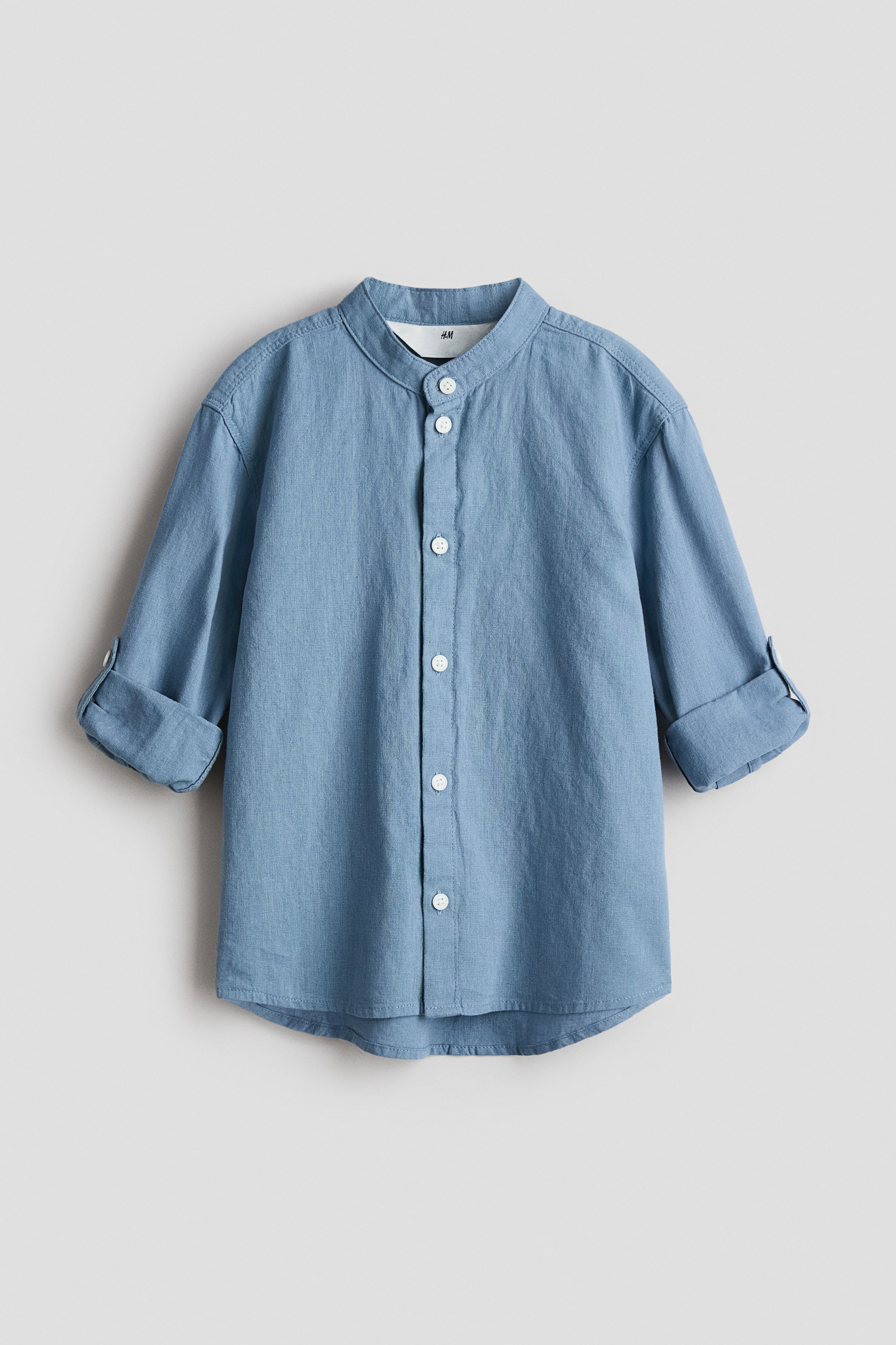 View larger image: A light blue Grandad shirt is presented FRONT, featuring a band collar and a front placket with white buttons. Its long sleeves are rolled up and secured with a tab and button, and it is displaying a soft, airy weave and a gently rounded hem.