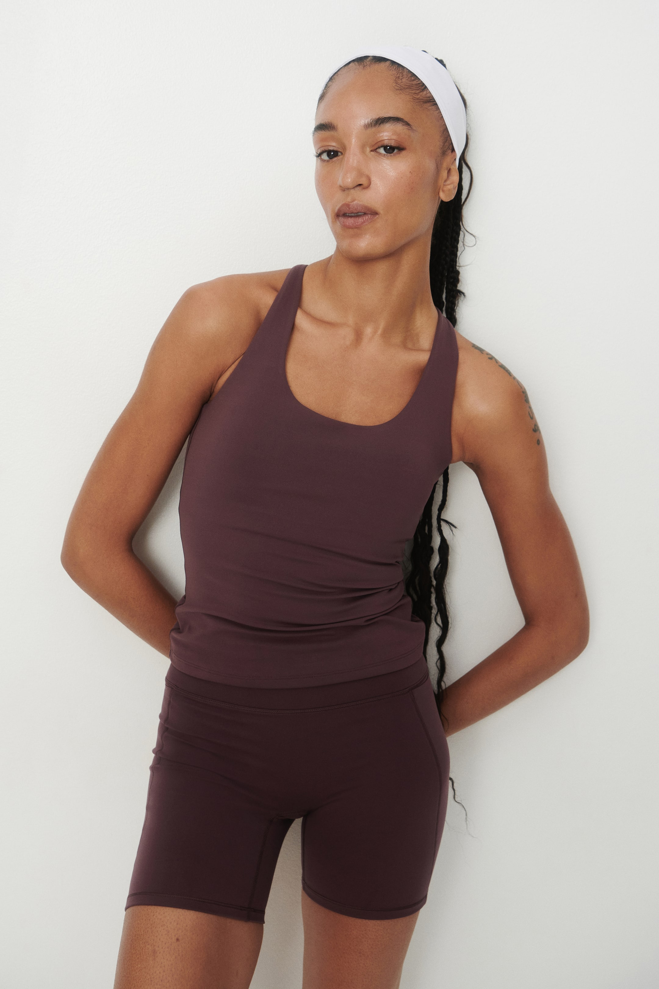 Grotere afbeelding bekijken: Een donkerpruimkleurige activewear-set, bestaande uit een getailleerde tanktop met een ronde hals en een bijpassende high-waisted fietsbroek, wordt gedragen door het model. De sportieve outfit is gestyled met een eenvoudige witte haarband.