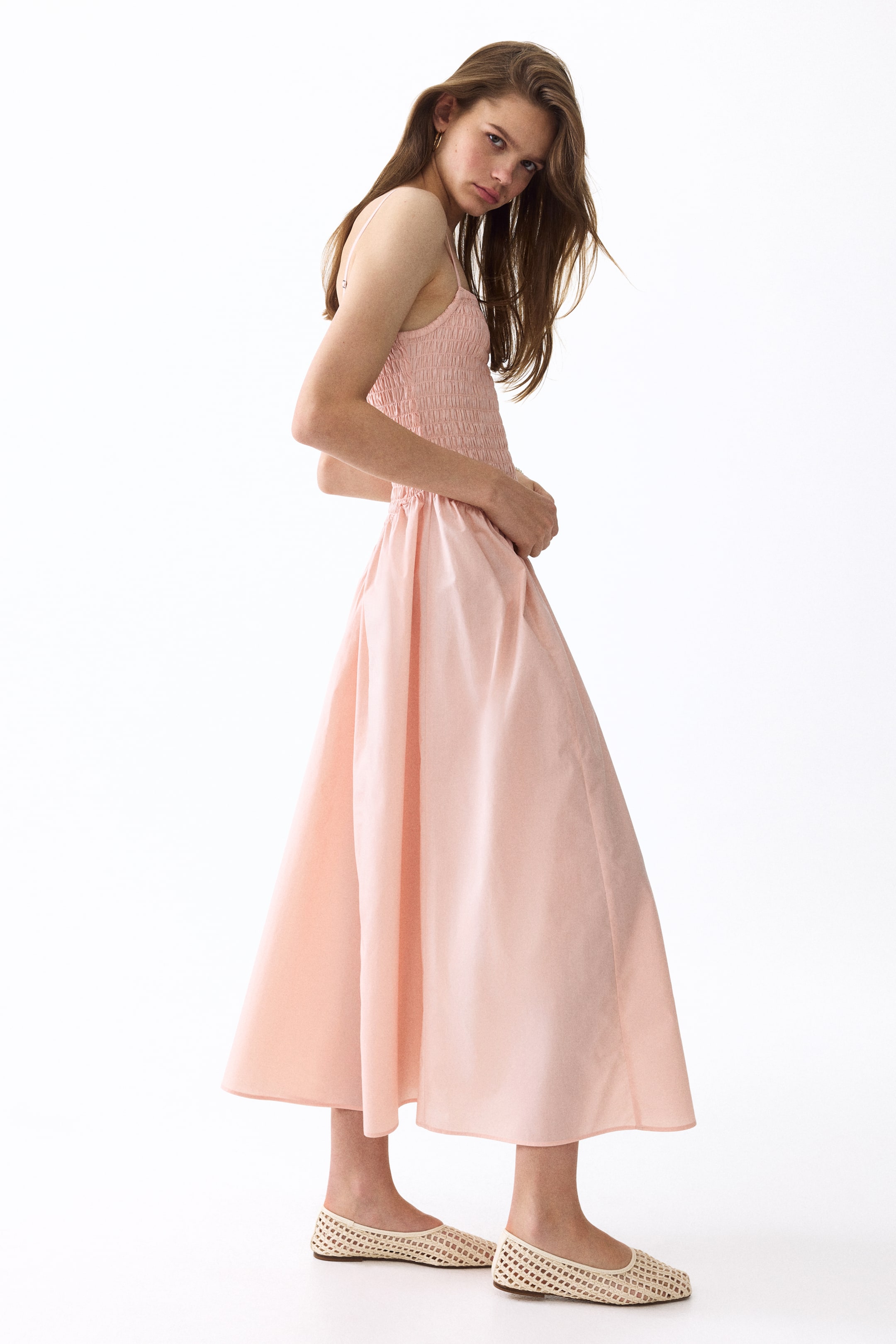 View larger image: The model is wearing a pink maxi dress crafted from an airy cotton weave. This piece showcases a straight cut at the top, a smocked bodice with slender shoulder straps, a V-shaped gathered seam at the waist, and a flared skirt. The model is positioning her hands on the dress at the waist and is paired with light-coloured woven flat shoes.