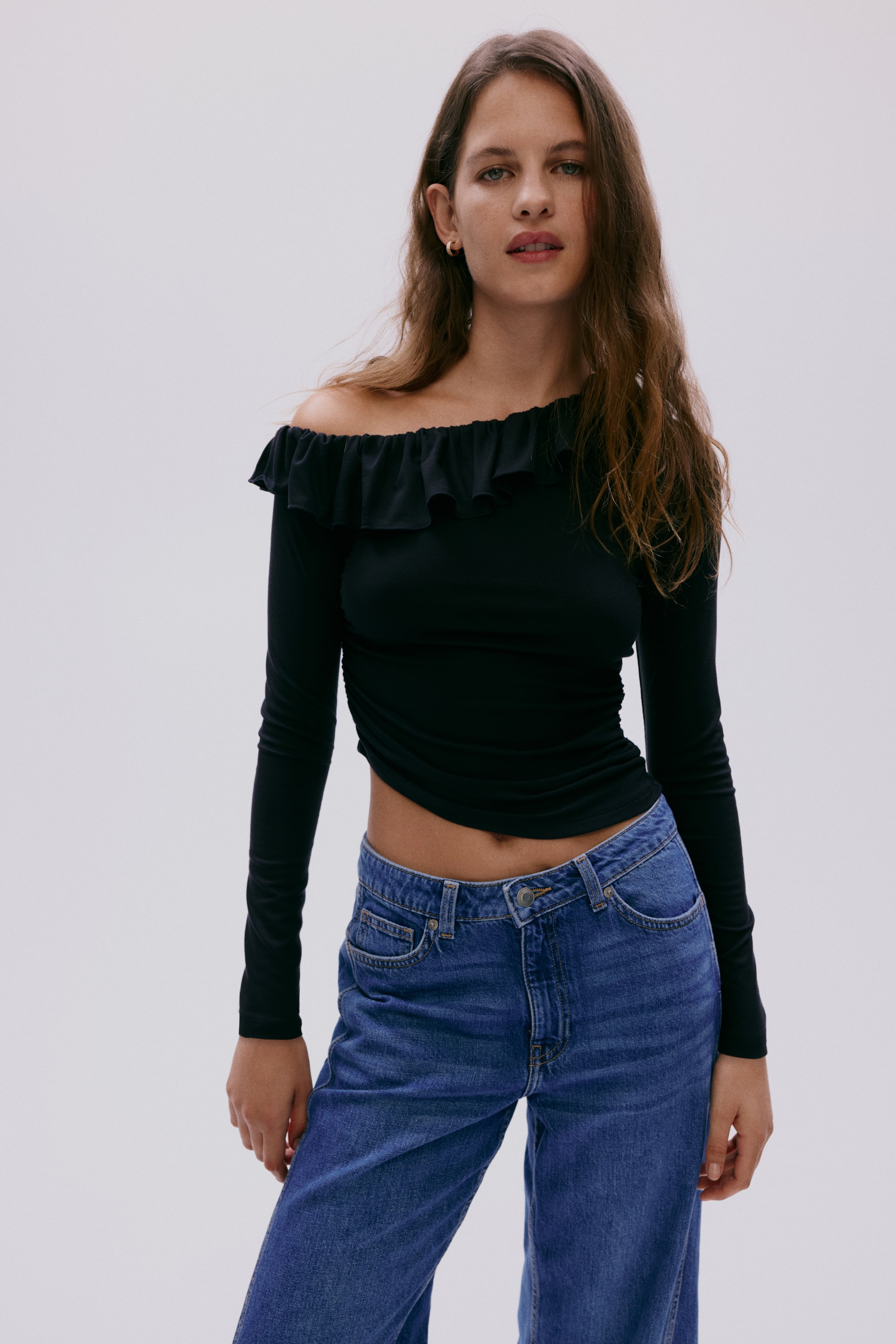 View larger image: This black long-sleeved top showcases an asymmetrical ruffled off-shoulder neckline, revealing the right shoulder. It highlights a fitted silhouette with subtle side ruching and a cropped hem, paired perfectly with blue jeans.